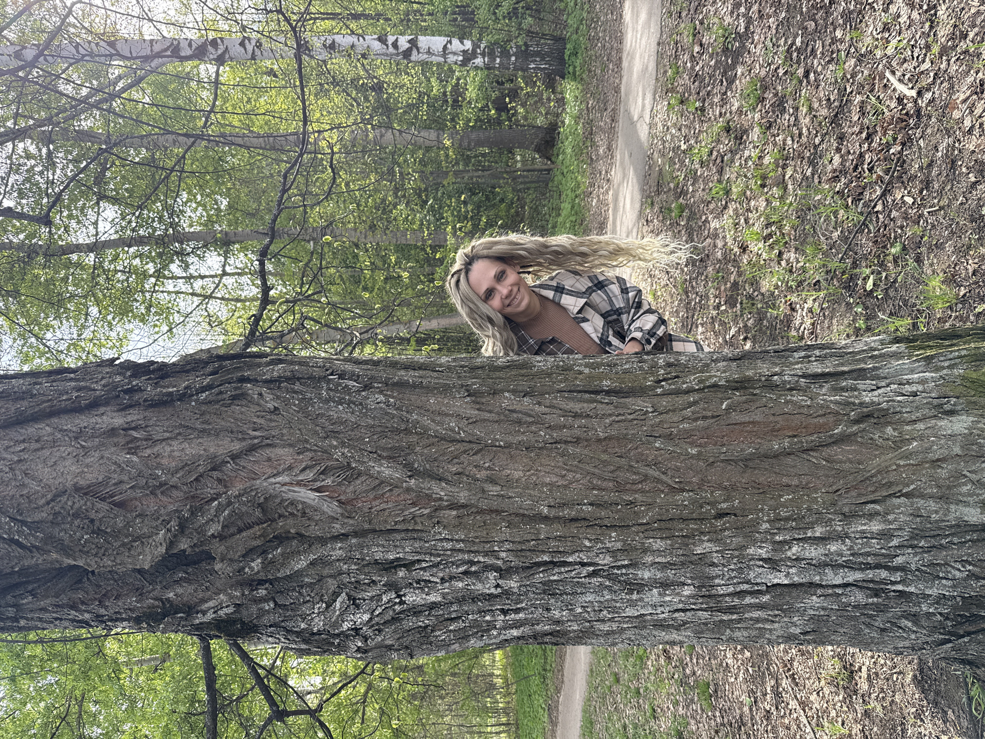 The image shows a woman with long blonde hair, wearing a plaid shirt, standing behind a large tree trunk in a park or wooded area. The tree dominates the foreground, with the woman's face and upper body visible behind it. A path is visible to the right, covered with leaves. A forest with green foliage can be seen in the background. The location is Rusanovo, Russia.