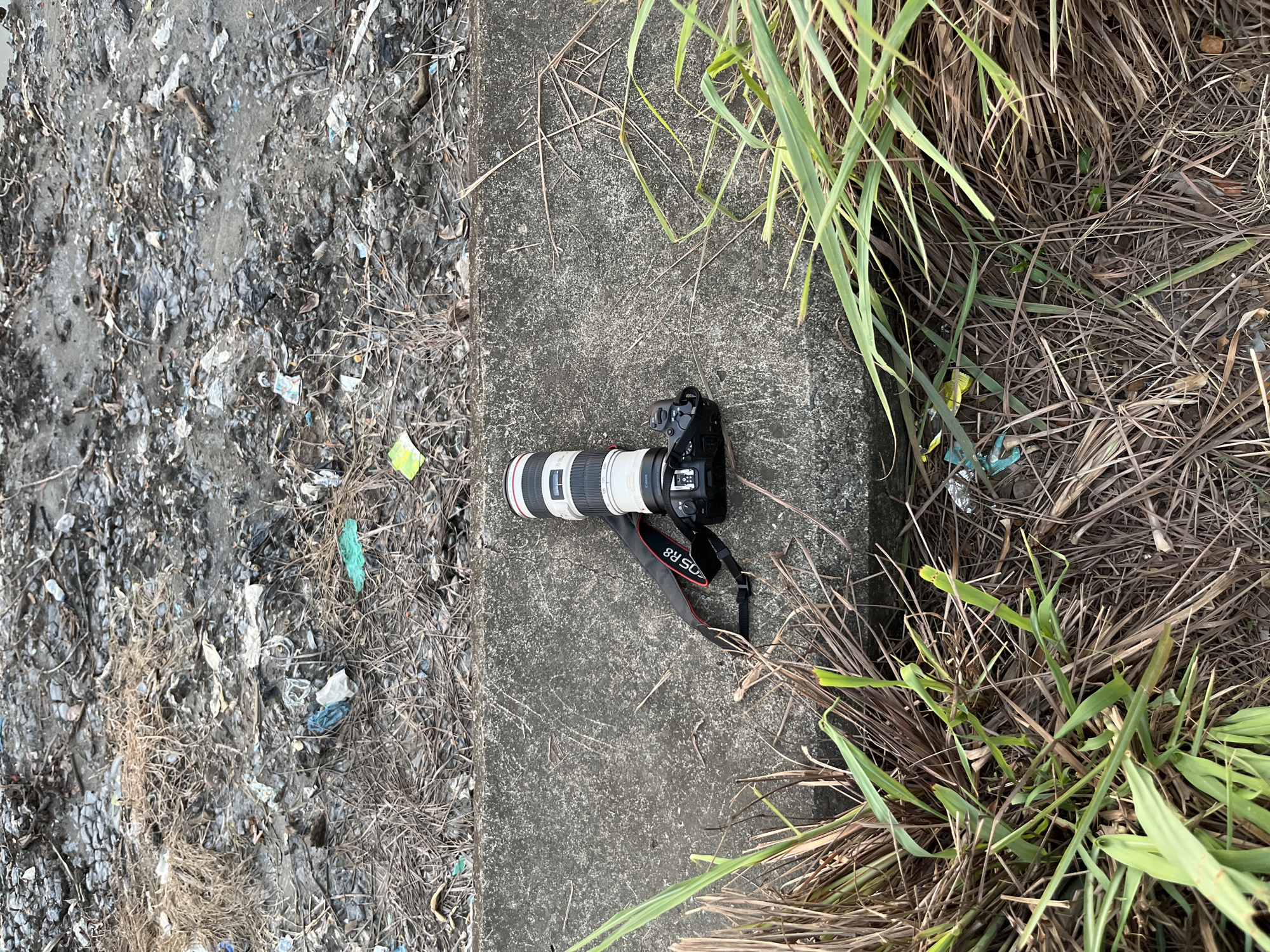 A DSLR camera with a telephoto lens attached rests on a concrete ledge. The camera has a black body and a white lens with red and black accents. A black and red strap is attached to the camera. The concrete ledge is bordered by dry grass and debris on one side and more dry grass and debris on the other. The background consists of a patch of dry ground with scattered litter. This scene is located in Lhokseumawe, Indonesia, on a dirt roadside.
