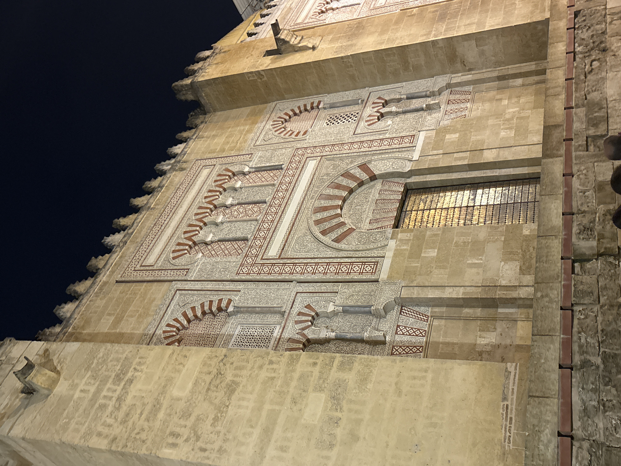 The image depicts a portion of the exterior wall of the Mezquita-Cathedral of Córdoba, Spain, photographed at night. The wall is constructed of light-colored stone, displaying intricate geometric and arabesque designs. The patterns are accented with red and white details. A barred window is visible, glowing with interior light. The sky is dark in the upper left, and the stone wall shows weathering and variations in color, hinting at its age.