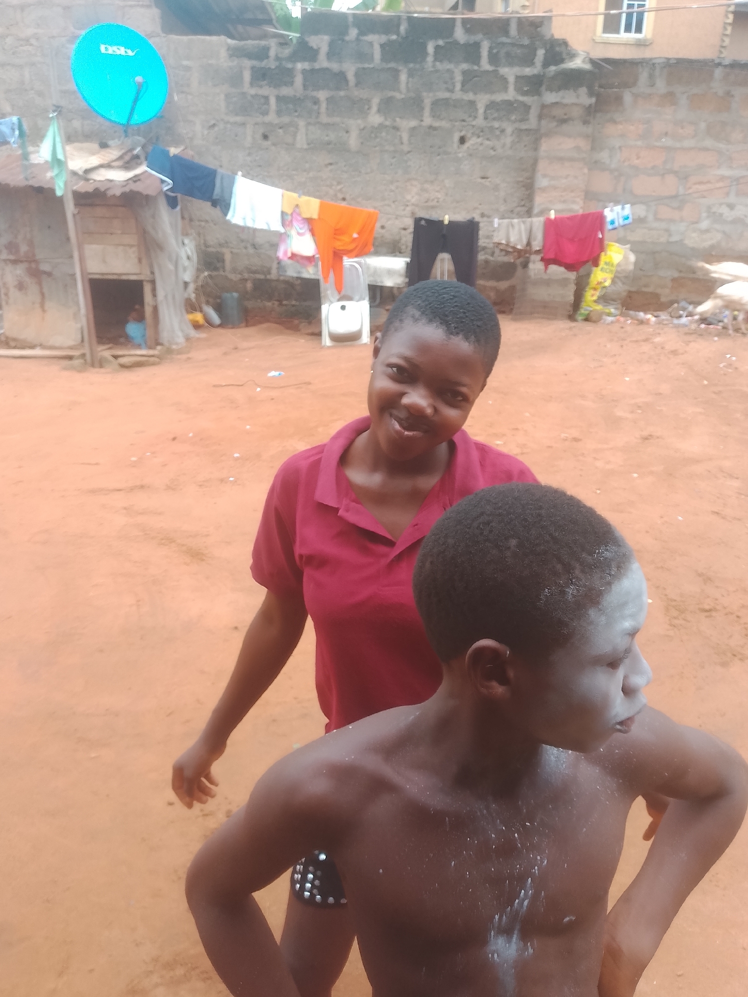 In an outdoor scene in an unspecified location in Unknown, Unknown, a young woman in a maroon collared shirt smiles at the camera while standing behind a shirtless young man. The young man has what appears to be white powder scattered on his torso and face. In the background, clothes are hanging on a line strung between a brick building and a rough shack with a turquoise satellite dish affixed to it. The ground is dirt.
