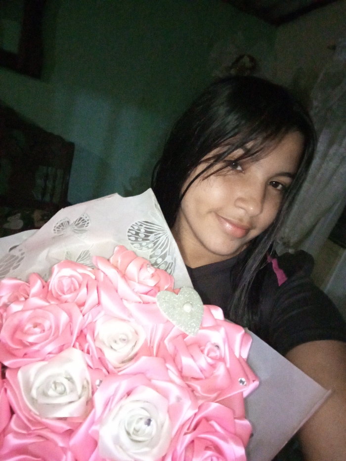A young woman with dark hair smiles slightly while holding a bouquet of pink and white satin ribbon roses wrapped in white paper. The roses feature small rhinestones and a glittery heart embellishment. The woman wears a black t-shirt and has light skin. The background is a dimly lit room with a dark green wall and a glimpse of furniture.