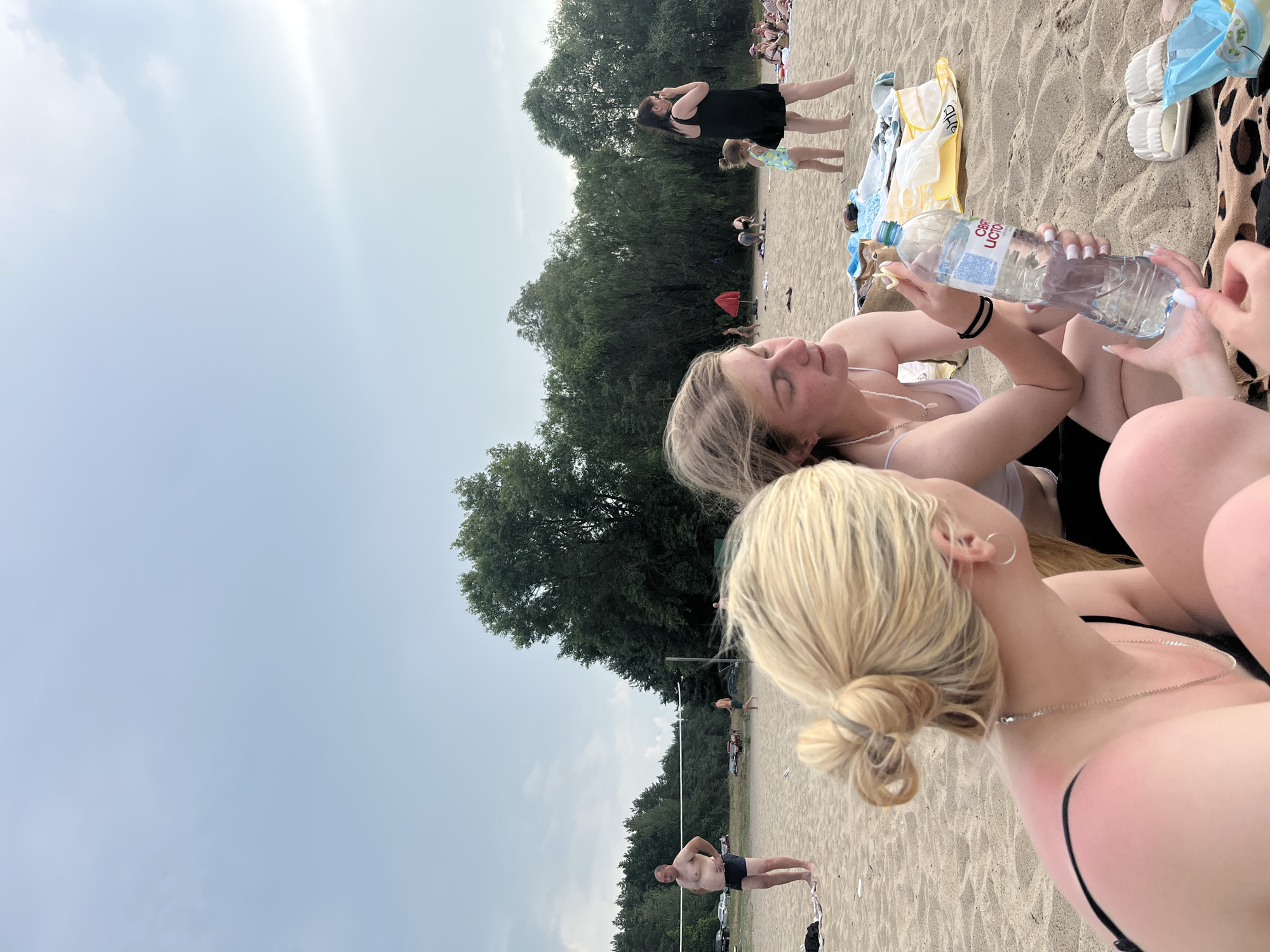 This image depicts a sunny day at a beach, likely in Belarus. The primary subjects are a group of young women enjoying leisure time on the sand, with a body of water or lake visible in the background. The atmosphere is relaxed and summery, with people casually dressed in swimwear.
In the foreground, two young women are prominently featured. One is holding a clear plastic bottle of water, and her nails are painted with white polish. The other woman is lying down, with her eyes closed, appearing to bask in the sun. The presence of other beachgoers in the background, some sitting and others standing, suggests a busy public beach.
Further back, trees line the edge of the beach, indicating a natural, possibly forested area surrounding the location. The sky is a bright, overcast blue, suggesting it is daytime and the weather is warm, though potentially with some cloud cover. Some beach accessories like towels and sandals are visible scattered on the sand. The overall scene evokes a sense of summer vacation and outdoor recreation.
There is visible text on the water bottle which appears to be in Cyrillic script, "ВИТАМИННО-МИ_ЕРАЛ". The image has been rotated 90 degrees counter-clockwise.