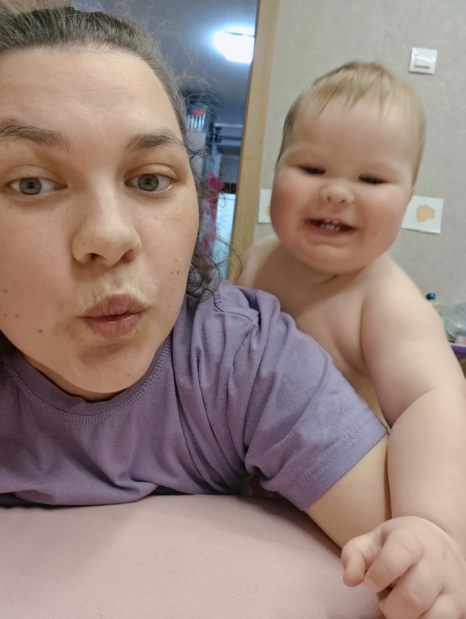 A woman wearing a purple t-shirt is holding a baby. The baby is shirtless and smiling. The photo appears to be taken indoors. The background includes a doorway and a wall with some decorations. The location is in Chaikovsky, Russia.