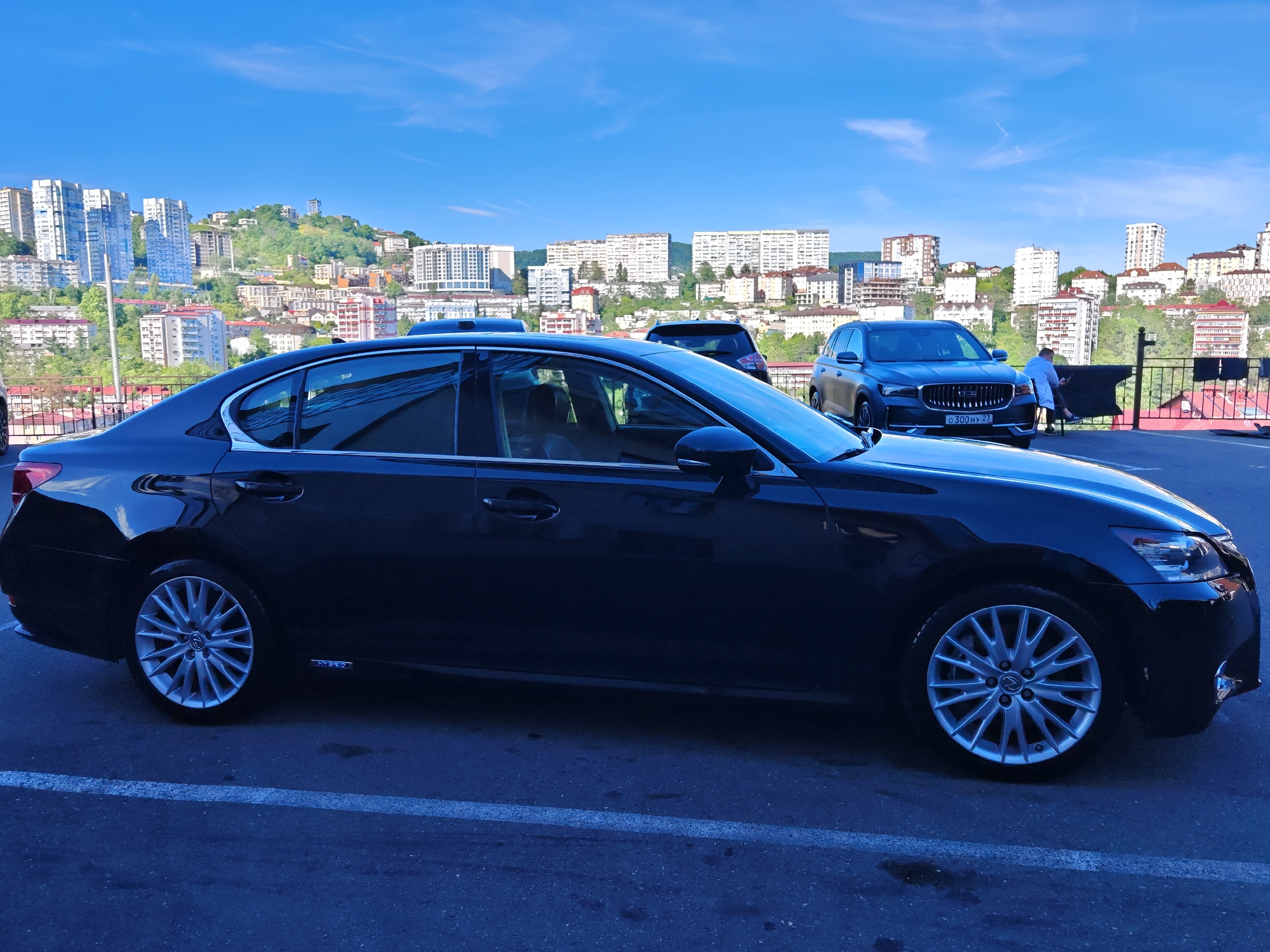 The image captures an urban scene in Sochi, Russia, under a bright blue sky with sparse white clouds, indicating daytime and clear weather.
In the foreground, a dark-colored Lexus sedan, likely a GS model, is prominently parked on an asphalt surface, partially visible. A "HYBRID" badge is visible on its side, and it features distinctive multi-spoke alloy wheels. To the right and slightly behind the Lexus, a gray Geely SUV, possibly a Monjaro or Tugella model, is also parked. Its front license plate displays "O 300 HY 23"; the "23" region code confirms its location within Krasnodar Krai, where Sochi is situated.
Adjacent to the gray SUV, a person, appearing to be a man in a light-colored shirt or uniform, is seated on a black chair next to a dark metal fence. He seems to be resting or engaged in an activity on a device, looking towards the right. Fabric or covers are draped over the fence behind him.
The background presents a vibrant cityscape built on rolling hills. Numerous multi-story residential and commercial buildings, ranging from modern high-rises with light facades to older structures, are densely packed across the landscape. Patches of green vegetation are visible among the buildings, highlighting the hilly topography characteristic of Sochi. The scene suggests a typical day in a bustling coastal city.