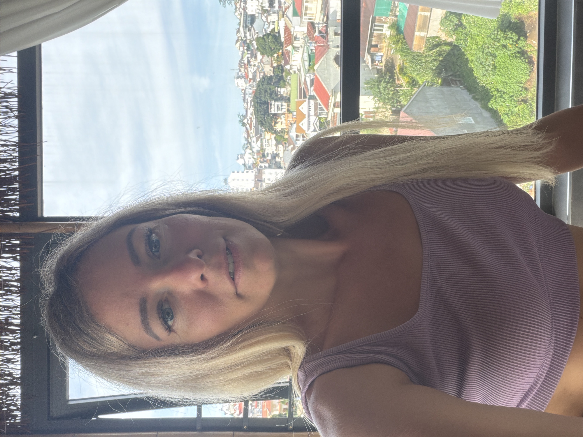 A light-skinned woman with blonde hair is looking at the camera with a neutral expression, her mouth slightly open. She is wearing a light purple ribbed tank top. She is indoors, with a thatched roof structure above her and a window to her right with a white curtain. The window overlooks a densely populated cityscape with a variety of buildings, many with red or orange roofs, and lush green trees. This appears to be the city of Da Lat, Vietnam, known for its hilly terrain and European-style architecture. The lighting suggests it is daytime, possibly late morning or early afternoon.