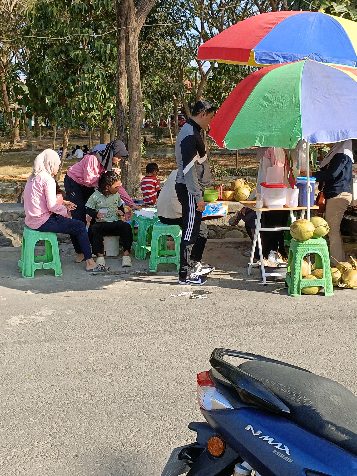 The image depicts a street scene in Bulukumba, Indonesia, with a group of people gathered around a roadside stall selling coconuts. The stall is shaded by colorful umbrellas, a red and blue one above a green and blue one. A man in a black and grey jacket and black pants stands near the stall, facing away from the camera. To his left, a woman in a pink hijab and pink top sits on a green plastic stool, while another woman in a black hijab and pink top leans over to interact with a young person sitting on another green stool. This young person, wearing a green shirt and black pants, is holding a straw and appears to be drinking from a cup. Several green plastic stools are visible around the stall. Piles of coconuts are displayed on the ground and on a green stool. In the background, trees and some parked vehicles are visible. The foreground is dominated by the asphalt road, with a portion of a blue scooter visible at the bottom of the frame.
