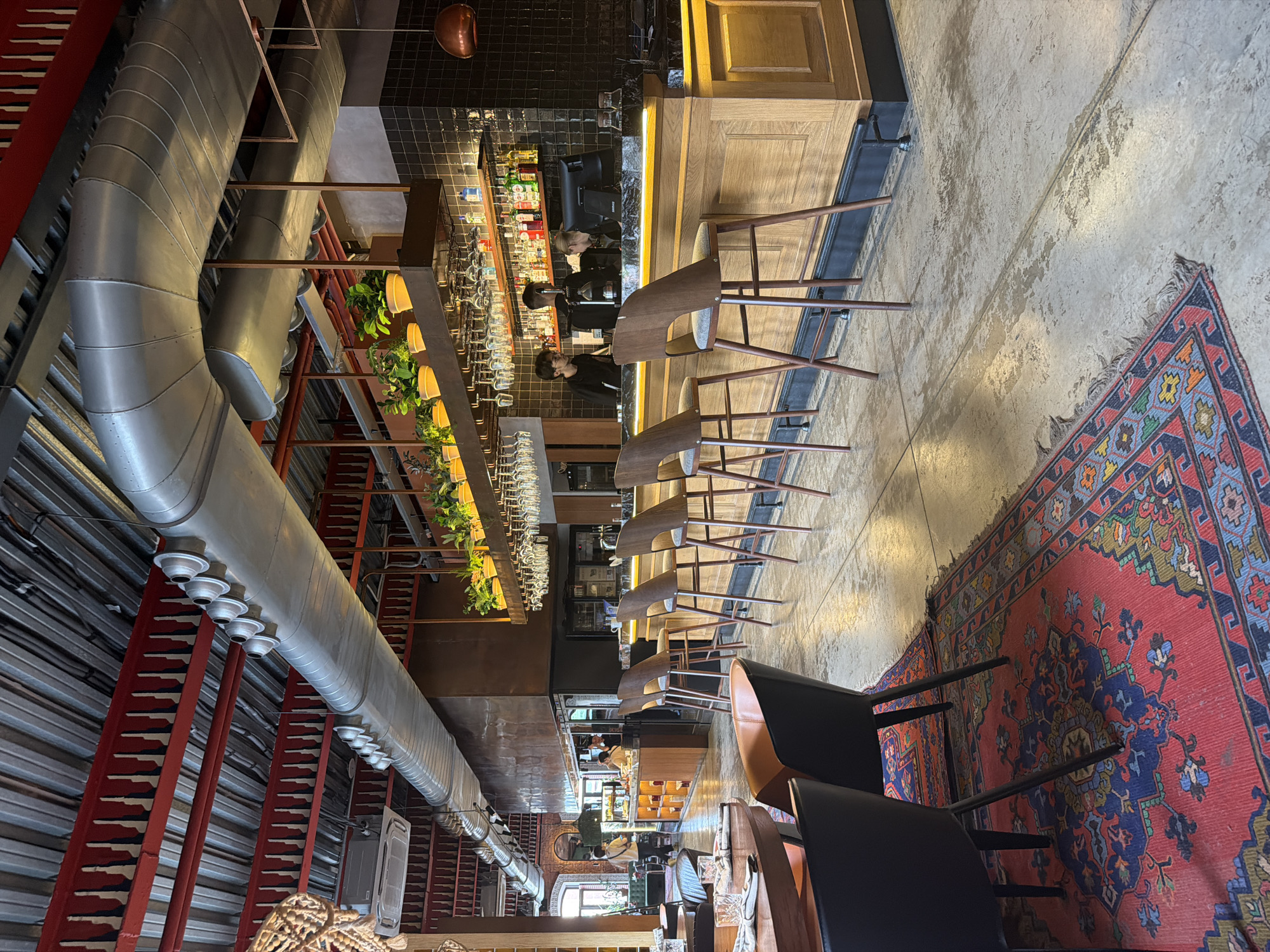 An interior shot of a modern restaurant or bar shows a long wooden bar with stools in the foreground, tables and chairs to the left, and a bar area with employees in the background. The ceiling features exposed metal ductwork and red structural beams. A colorful Persian rug is laid out on the concrete floor in front of the tables. Bottles of alcohol and glassware are visible behind the bar. The lighting is ambient, with some spotlights illuminating the bar area.