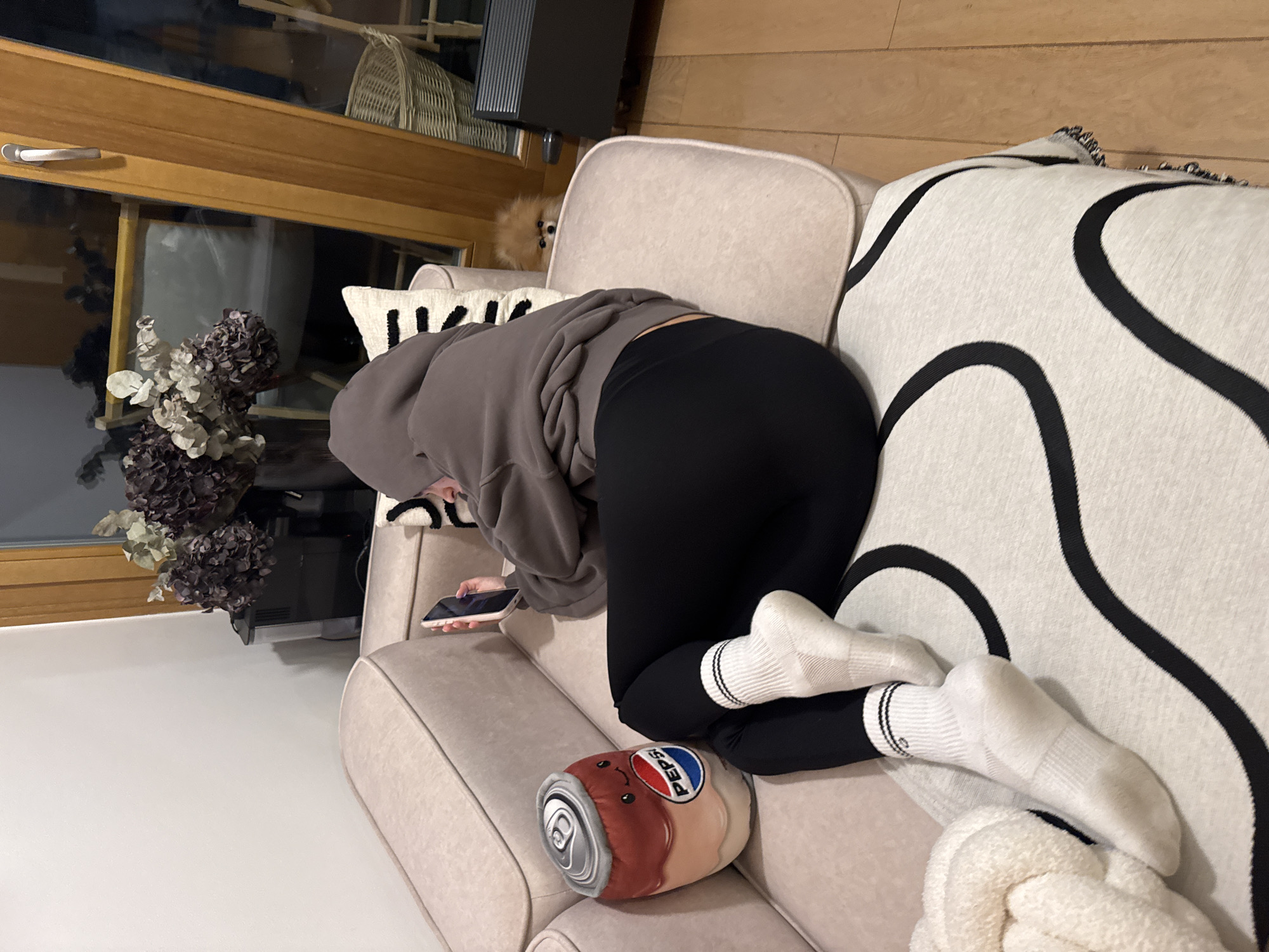 A woman wearing a gray hoodie and black leggings is on a beige sofa with white and black patterned pillows, holding a phone. Her feet are in white socks. A soft, Pepsi-can shaped pillow is next to her. A small dog is partially visible behind the couch. A window is in the background with a floral arrangement on the ledge. The location is in an indoor setting.