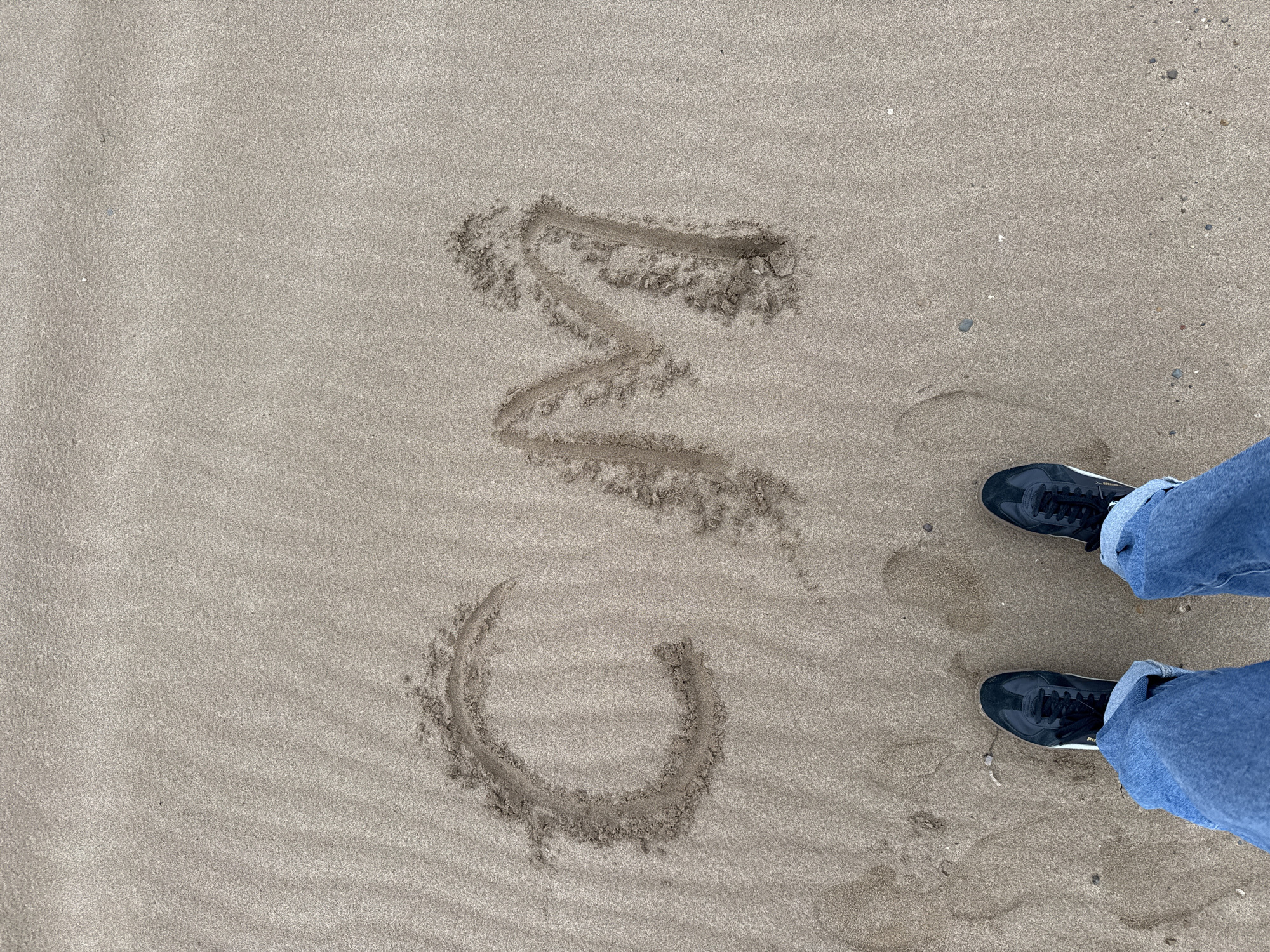 An overhead shot captures a sandy surface with a person's feet and written text. The sand is light greyish-brown, with some areas appearing slightly darker, suggesting dampness. Small, dark pebbles are scattered across the sand. The feet, clad in blue denim jeans rolled up at the ankles and black sneakers with white soles, are positioned towards the bottom right of the frame, pointing slightly inwards. The sneakers have black laces, and a faint brand name, possibly "PUMA," is visible on the side of one shoe. In the upper left and center of the sand, the letters "OMG" are clearly written, etched into the sand, with small piles of displaced sand forming the outlines of the letters. The scene is located in Noviy, Russia.