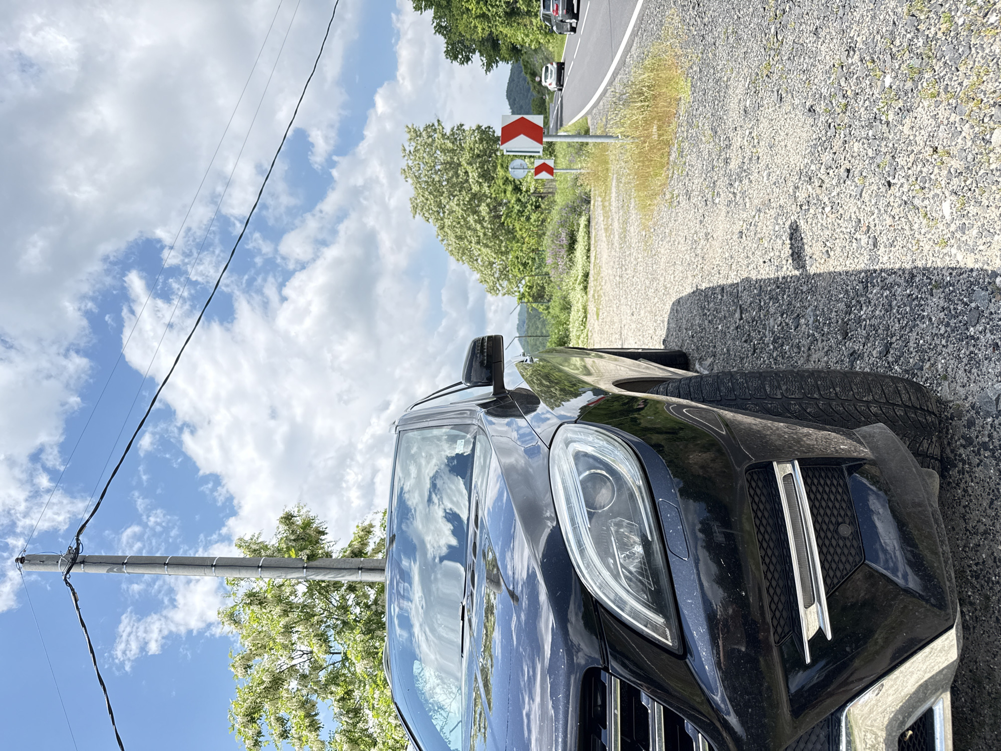 The image depicts a roadside scene in Chubrika, Bulgaria, featuring a black Mercedes-Benz ML-Class SUV parked on a gravel shoulder. The vehicle's front left quarter is prominent in the foreground, showing a dusty surface and reflections of a partly cloudy sky on its hood and windshield. To the right of the parked vehicle, a two-lane asphalt road with a solid white shoulder line and a dashed white center line curves sharply to the right. Two red and white chevron road signs, indicating a curve to the right, are visible on the roadside. Two light-colored passenger cars are observed traveling in the distance along the road. In the upper left, a concrete utility pole supports multiple overhead power lines against a sky with scattered white clouds and patches of blue. The surrounding environment includes various trees, notably one with white blossoms, along with green foliage, bushes, and sparse grass. Shadows cast by the SUV indicate sunlight from the left. No individuals are present in the scene.