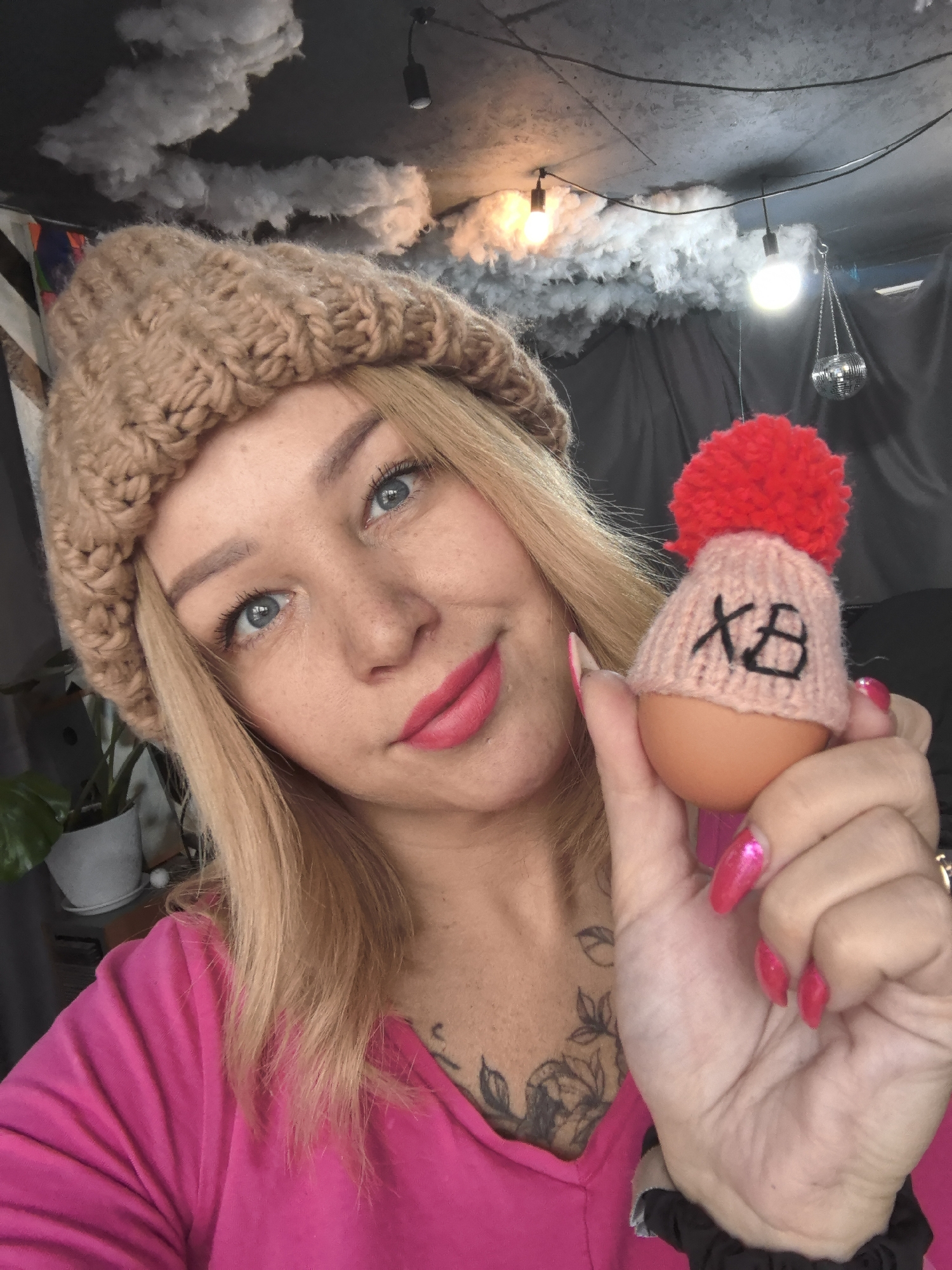 A woman with blonde hair and blue eyes, wearing a tan knitted hat and a pink shirt, holds an egg in her right hand. The egg is wearing a small pink knitted hat with a red pompom and the letters "XB" written on it. The woman has freckles and is wearing pink lipstick. Her nails are painted pink. She has a tattoo on her chest. The background includes faux clouds and string lights, along with a disco ball and a plant. The specific location within the city of Noviy, Russia is not discernible from the image.