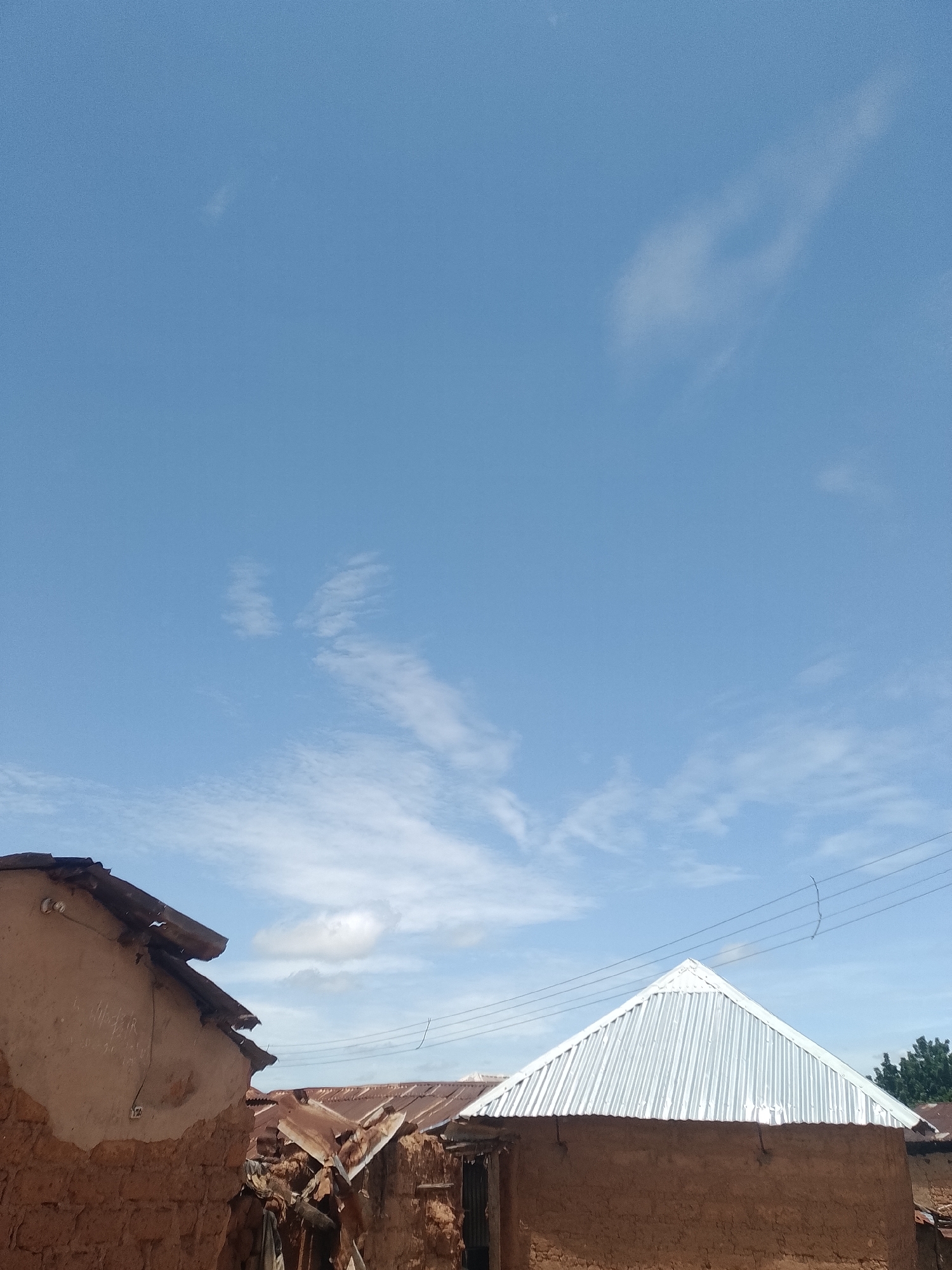 A low-angle, eye-level, outdoor, daytime, full shot shows a bright blue sky with wispy white clouds above several structures with rustic, mud-built walls and corrugated metal roofs. The structure in the foreground left has a damaged, brown, corrugated metal roof with visible rust. A portion of a white electrical conduit is visible on its exterior wall. To the right, a building with a conical, silver, corrugated metal roof is centered in the frame. Further to the right, another building with a brown, corrugated metal roof is visible. Electrical wires are strung across the scene above the buildings. The environment appears to be a rural or semi-rural area.