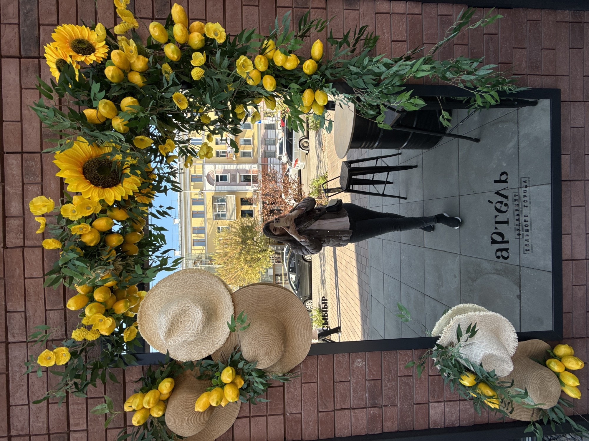 A vertically oriented photograph captures a scene outside a building with a red brick facade. A rectangular mirror framed in black reflects the surrounding environment. Yellow flowers, sunflowers, green foliage, and straw hats are arranged around the mirror, adorning the brick wall.
In the mirror's reflection, a person stands taking a photo with their phone. The reflection also shows a street with cars, buildings, trees, and a gray tiled sidewalk. A black table and chairs are visible in the reflection.
The mirror has the word "артель" written on it, along with the phrase "ФУДМАРКЕТ ЭТО ГОРОДА". This is a reference to Artel Food Market in Krasnoyarsk, Russia.