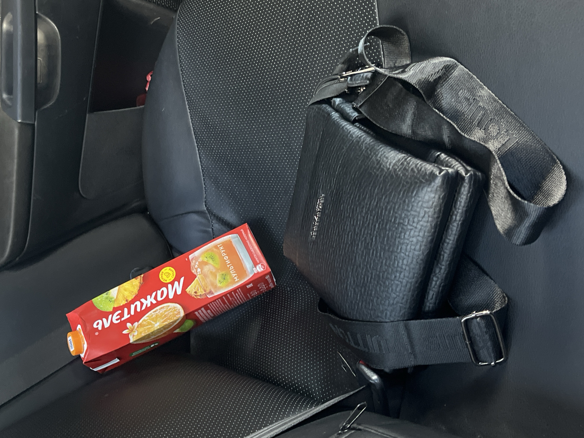 The image depicts a black leather satchel and a carton of "Мохито" juice placed on the perforated leather seat of a car, likely in Kostroma, Russia. The satchel is positioned in the foreground, with its strap partially covering the seat. The juice carton, red with fruit imagery and text, is situated behind and to the left of the satchel. The car's interior, characterized by black leather seats and trim, suggests a modern vehicle. The lighting indicates it is daytime. No individuals are present in the frame, and there are no discernible actions or interactions occurring. The scene is static, focusing on the arrangement of the objects within the car.
