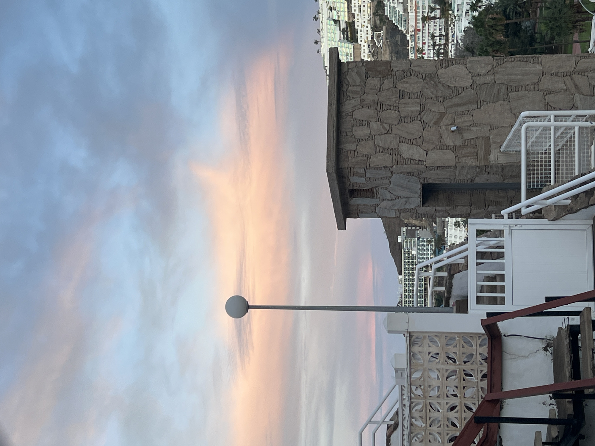 The image depicts an elevated outdoor scene in Mogán, Spain, at either sunrise or sunset, characterized by a sky displaying hues of pink, orange, blue, and grey. In the immediate foreground, a weathered set of stairs with dark reddish-brown metal supports and wooden treads is partially visible on the left. Adjacent to these stairs is a white metal gate with vertical slats, which appears to lead to another set of white-railed stairs descending out of view. To the left of the gate, a decorative concrete block wall with a circular pattern is present. A tall, slender grey pole with a spherical white lamp fixture on top stands prominently towards the left-center.
On the right side of the foreground, a robust, rectangular stone structure, possibly a small building or utility enclosure, is constructed from irregular, light brown and grey stacked stones. This structure features a dark, recessed rectangular opening on its visible side. Beyond this structure, and extending into the midground and background, a hillside is densely covered with numerous white and light-colored buildings, many featuring balconies and some with blue or green rooflines/accents, indicative of a resort or residential development. Clusters of green palm trees are interspersed among the buildings and in the lower valley areas. No individuals are visible within the frame.