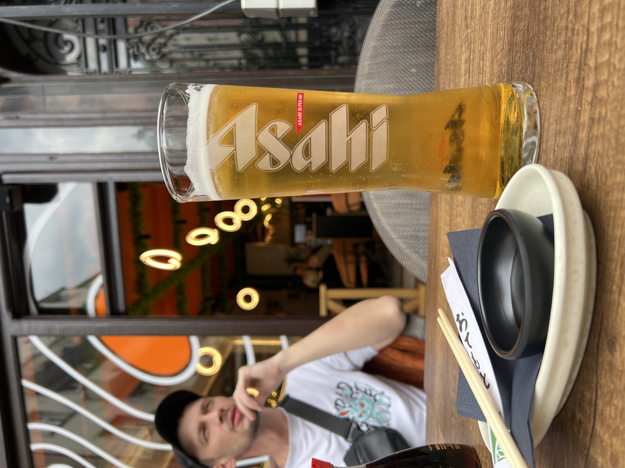 The image shows a glass of Asahi beer on a wooden table, next to a small white plate containing a black bowl, chopsticks, and a black napkin. A man is visible in the background, out of focus, wearing a white t-shirt and a black cap, with a strap across his chest. Through the glass doors and window, circular orange lights are visible, as well as other diners. Based on the visual elements of the image, it appears the location is a restaurant or bar in Belgrade, Serbia.