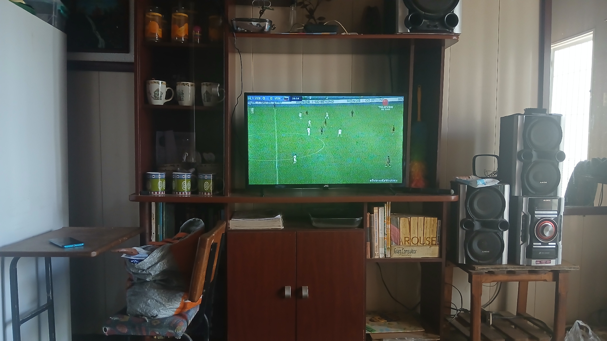 The image depicts a living room interior. A flat-screen television displays a soccer match between UZB and VEN, with the score 0-0 at the 39:54 mark. The TV is part of a large entertainment center with shelves filled with mugs, jars, books, and other decorative items. To the right of the entertainment center, a Sony stereo system sits on a small wooden table. A small folding table is on the left. A chair, partially obscured by bags, sits between the table and the entertainment center. Light is streaming in from a window on the right side of the frame. The OCR indicates the broadcast is from "TELEVEN" and mentions "#TelevenEsVinalinta", suggesting the image was taken in or relates to Venezuela. The text on the spine of the books on the shelf reads "Larousse Gran Consultor". Based on the OCR text on the television broadcast, the location is likely Altagracia, Venezuela.