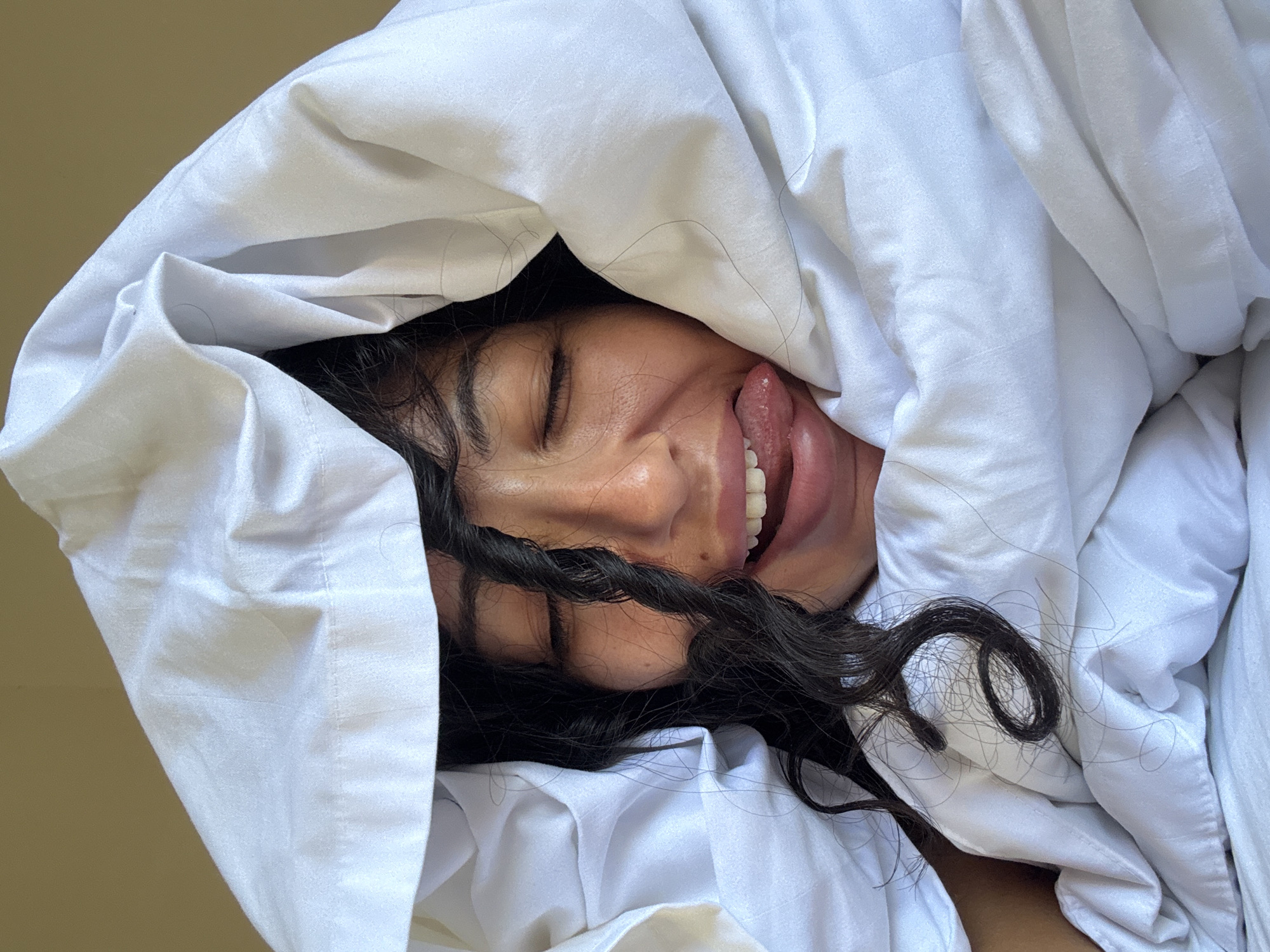 A close-up shot captures a person partially covered by a rumpled white blanket or duvet, lying down. The subject has long, dark, wavy to curly hair, parts of which are visible and some strands are draped over the white fabric. Their face is angled, with eyes closed and a playful expression, as they stick out their tongue. A few white teeth are visible as well. The skin tone appears warm.
The white bedding dominates the foreground and midground, with soft folds and shadows indicating its texture. In the background, a plain, light beige or light brown wall or surface is visible, suggesting an indoor setting. The lighting is soft and even, implying natural light, possibly daytime. The overall mood of the image is lighthearted and candid. No text is visible. Based on the provided context, the image was captured in North Kuta, Indonesia.