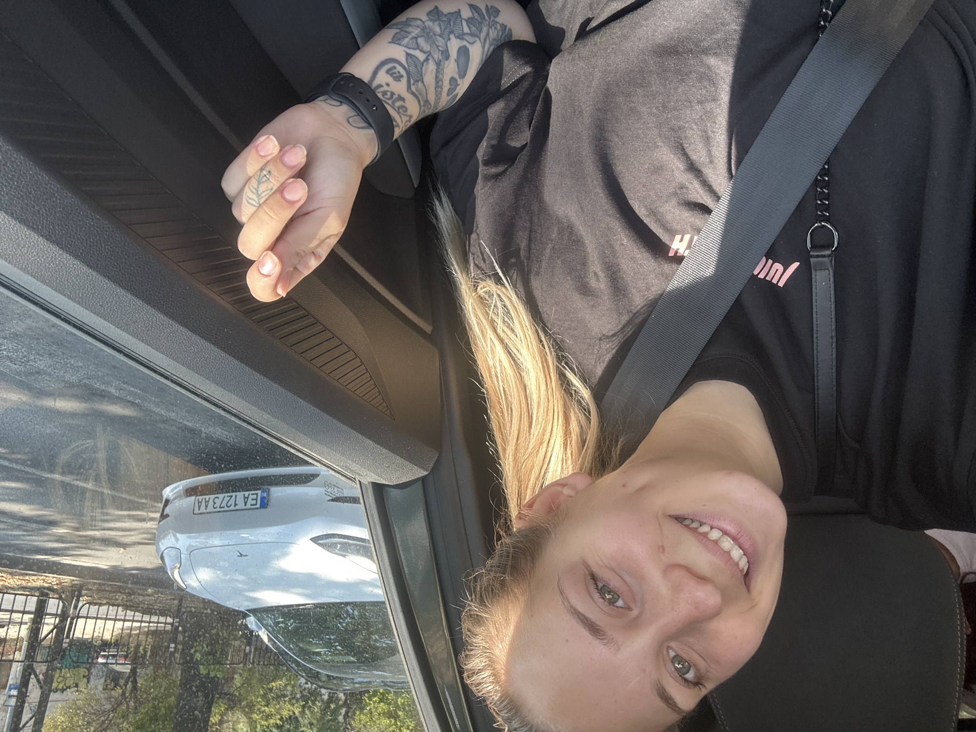 A young woman with blonde hair and green eyes smiles at the camera while sitting in the driver's seat of a car. She is wearing a black t-shirt with pink lettering and a black seatbelt is across her chest. Her left arm is visible, showing a black watch and several tattoos. Her right hand is raised, with her index finger and thumb touching. Outside the car window, a white Tesla Model 3 is visible, with a license plate that reads "EA 1273 AA". The car is parked on a street with trees and buildings in the background. The setting appears to be a sunny day in the city of Plovdiv, Bulgaria.