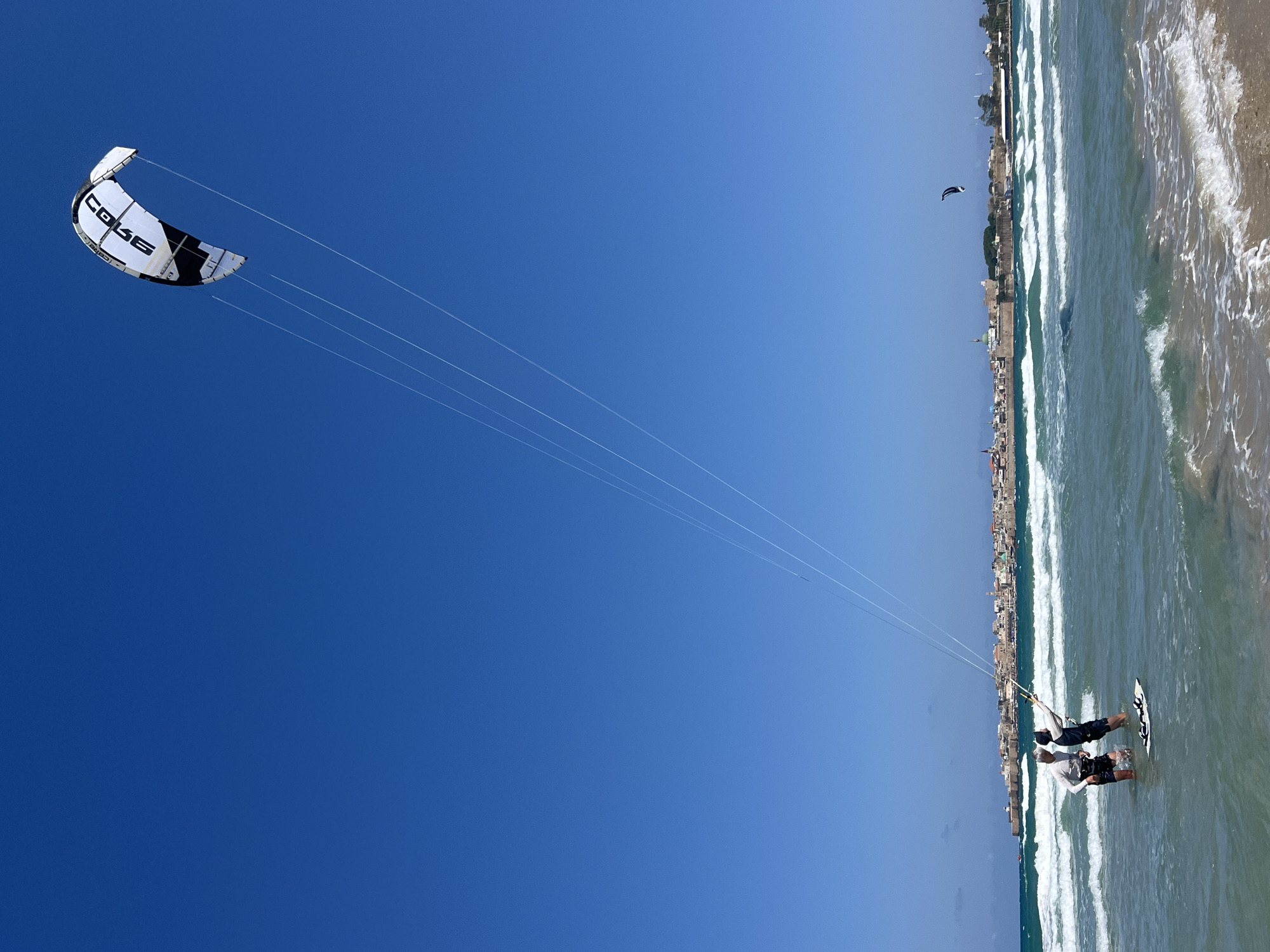 The image depicts a kiteboarding scene off the coast of Acre, Israel. In the foreground, two individuals are waist-deep in the water, one appearing to be preparing or assisting the other with the kiteboard. One person is holding the kiteboarding kite lines and kiteboard. The kite, a black and white Core model, is soaring in the sky. Along the coastline, a city skyline is visible. In the distance, another kiteboarder is seen in the water. The sky is clear and blue. The water is light turquoise and waves are breaking on the beach.