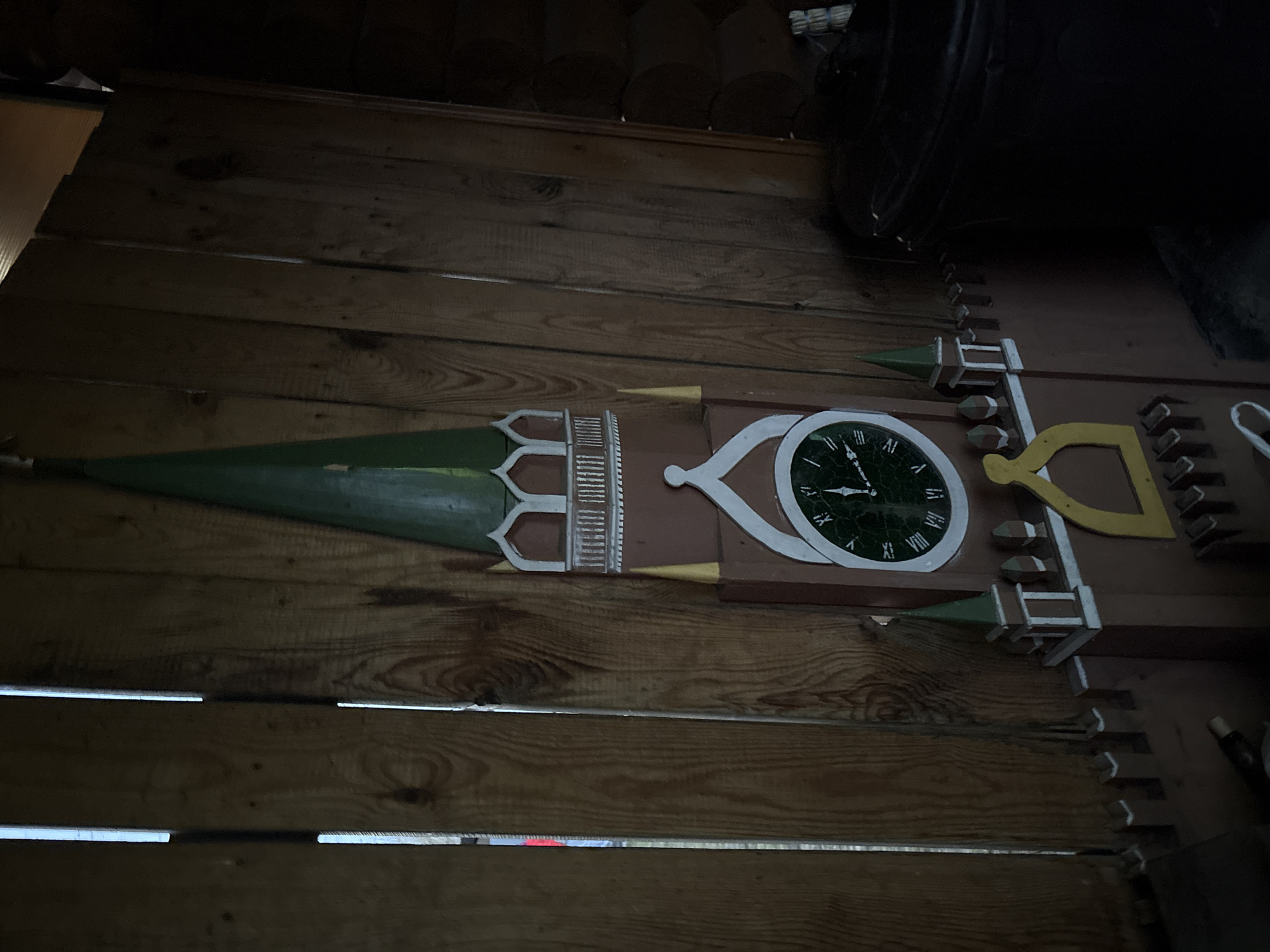 The image shows a wooden wall with a decorative, likely hand-crafted, wooden clock and architectural elements attached to it. The clock face is round, with Roman numerals, and appears to be made of green and white materials. A green spear-like ornament is attached to the left of the clock. The setting is dimly lit. The location is the city of Chaikovsky, Russia.