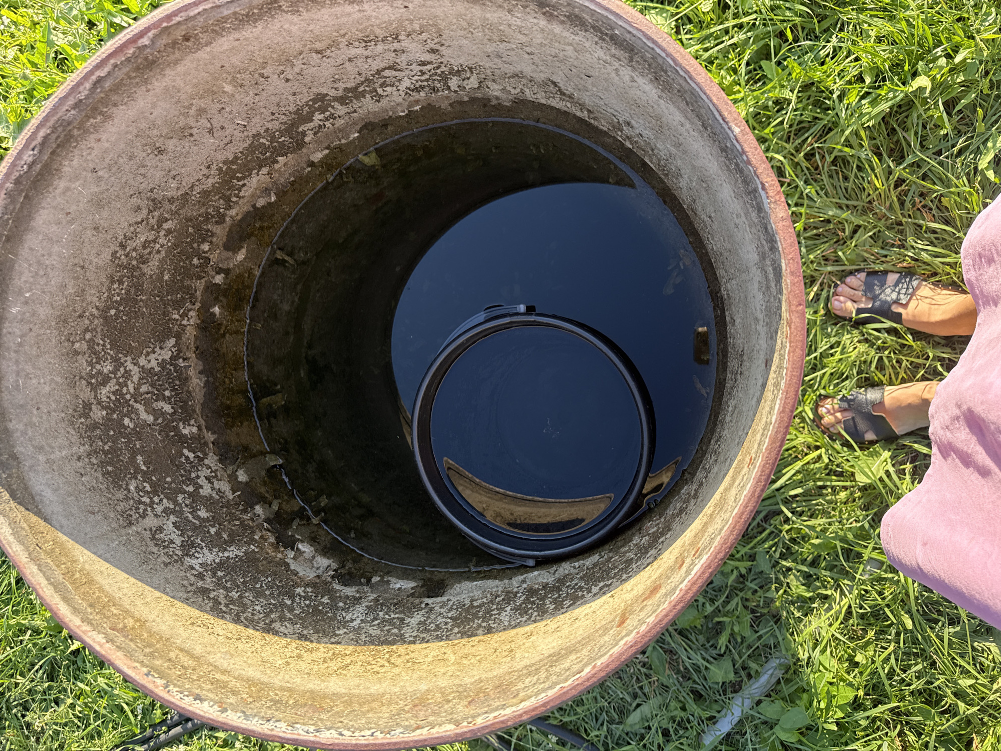 A first-person perspective shot looks down into an open, large, cylindrical concrete well filled with dark water. A black plastic bucket floats in the water. The inner rim of the well is weathered and discolored. The exterior of the well is partially visible, showing a reddish-brown rim. Green grass surrounds the well, and two bare feet wearing black sandals are visible in the foreground. The scene appears to be outdoors in daylight, possibly in Verkhneyarkeyevo, Russia, given the context provided.