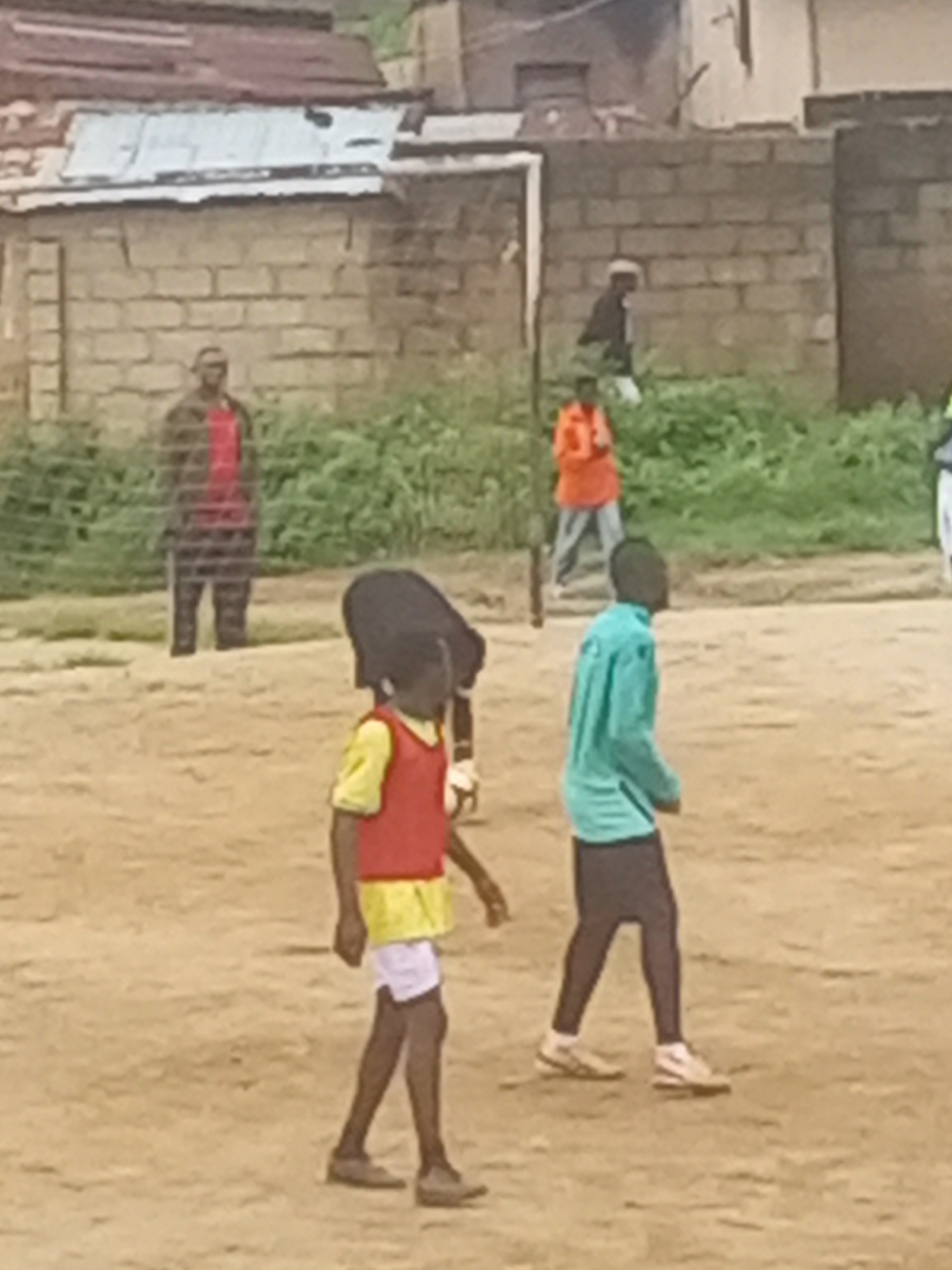 The image depicts a scene in Jos, Nigeria, with several individuals engaged in an activity on a dirt field. In the foreground, a young person wearing a yellow shirt and red bib is seen from the side, facing away from the camera. They appear to be holding or near a soccer ball. To their right, another person in a teal shirt and dark pants is also facing away, with their left arm slightly raised. In the background, a person in an orange shirt and light pants stands further back, and behind them, a person in a dark jacket and white head covering is partially visible. A man in a red shirt and dark pants is standing near a fence on the left side of the frame. The environment consists of a dusty ground, some vegetation, and buildings constructed from bricks and corrugated metal, suggesting an urban or semi-urban setting in Jos. The overall atmosphere appears to be casual, possibly related to a sporting event or a casual gathering.