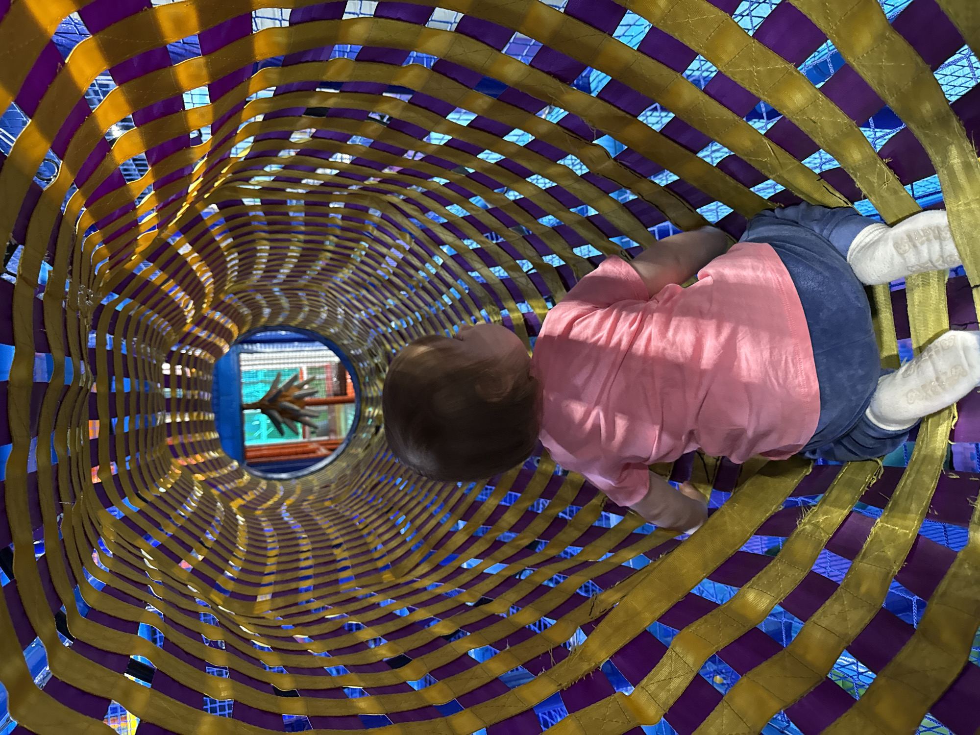 A child is crawling through a tunnel constructed of alternating gold and purple woven straps. The child is wearing a pink shirt, blue pants, and white socks. The end of the tunnel reveals another play area in the background. The location is Перовское сельское поселение, Ukraine.