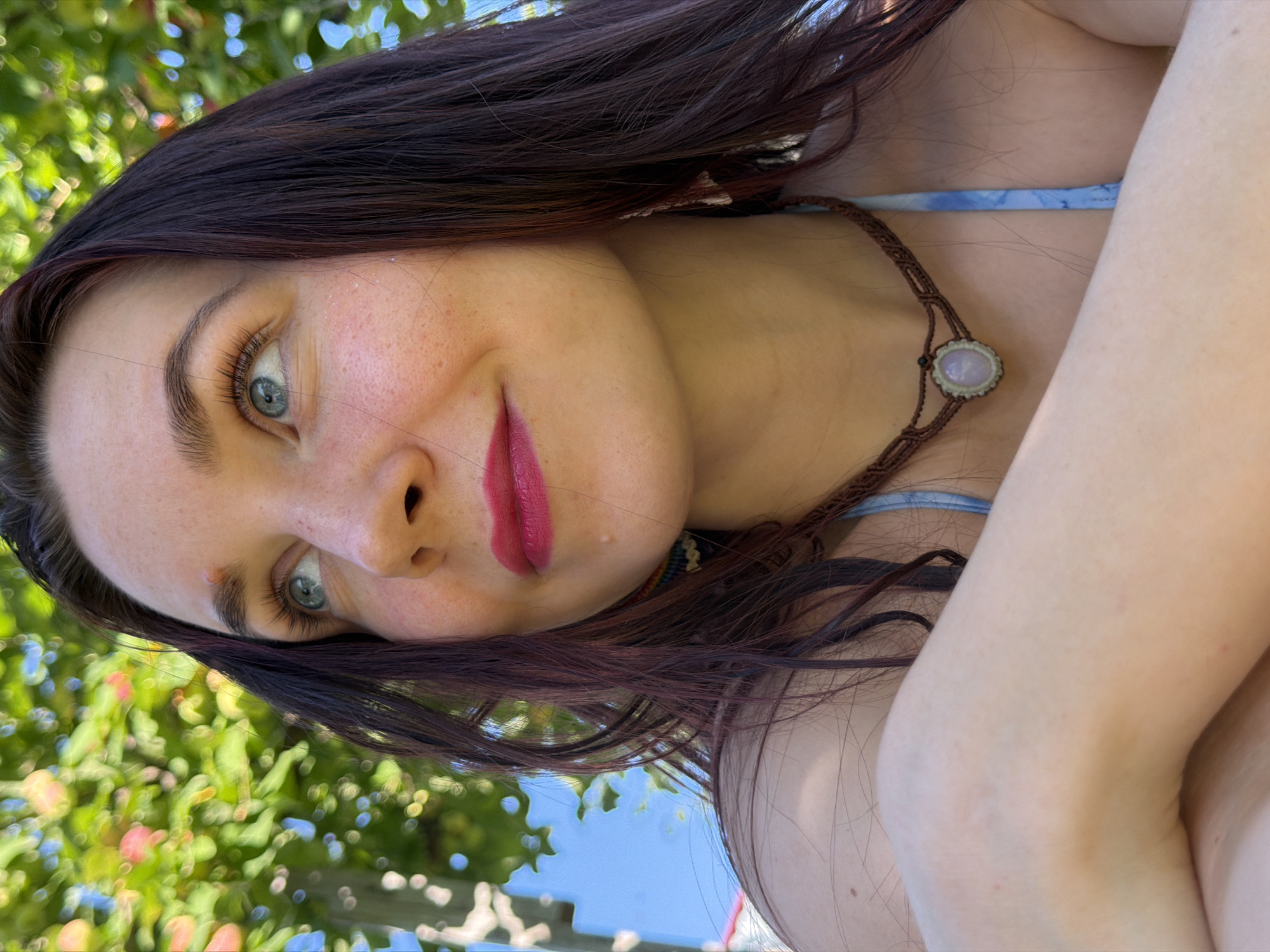This is a close-up, rotated selfie of a young woman outdoors. She has dark brown hair with auburn undertones, which frames her face. Her eyes are a striking blue-green, and she is wearing noticeable eye makeup and bright pink lipstick. She has a smattering of freckles across her cheeks and nose. She is wearing a light blue bikini top and a macrame necklace with a light-colored, oval-shaped stone pendant. Her skin appears fair.
The background is softly blurred, suggesting it is out of focus. On the left side of the image, lush green foliage with hints of pink and blue flowers is visible, indicating a natural setting with plant life. The upper right portion of the background shows a clear, light blue sky. The lighting suggests it is daytime, likely sunny, given the bright illumination and clear sky. The overall mood conveyed is casual and relaxed, possibly taken during warm weather. There is no visible text in the image.