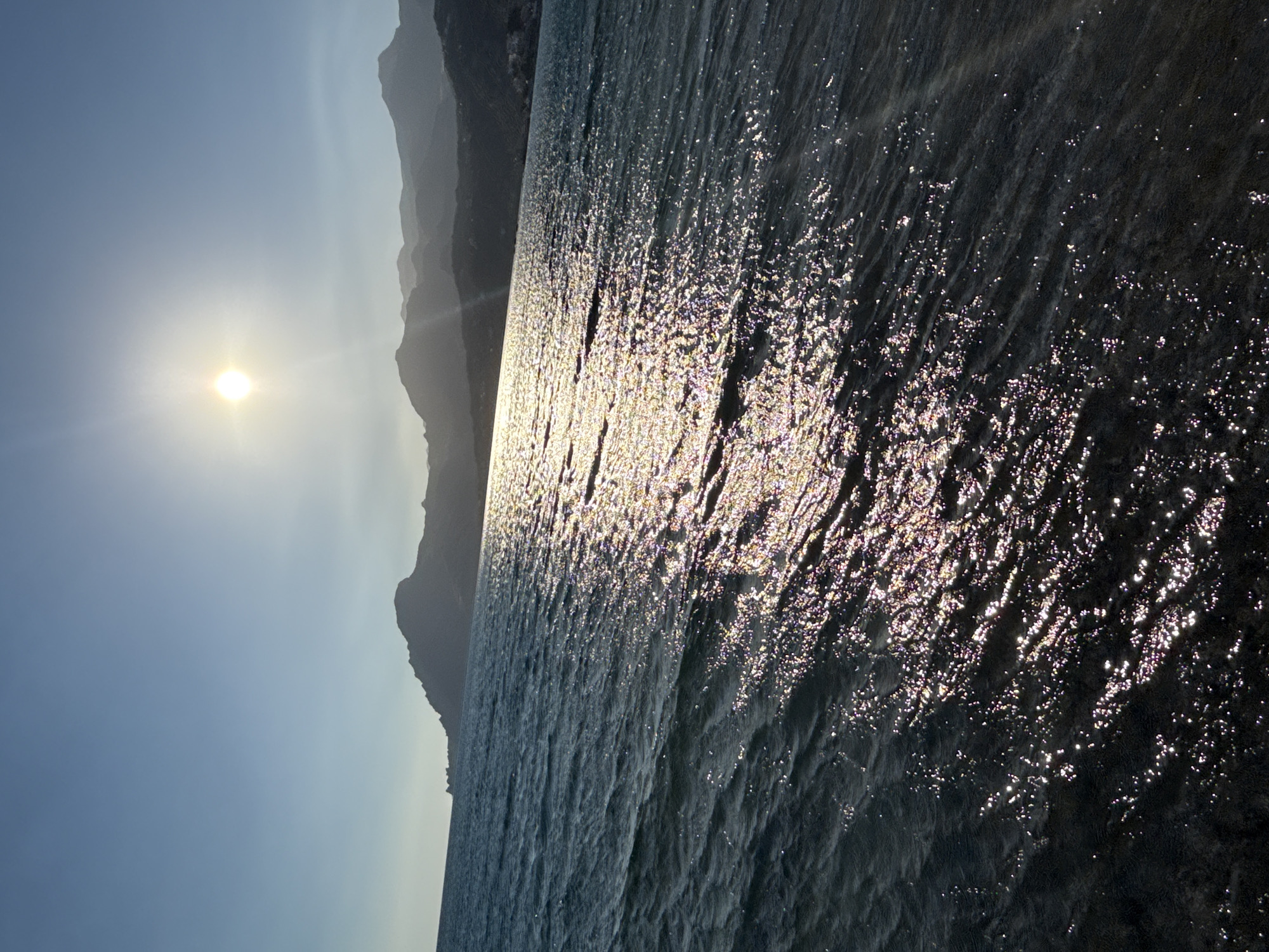 A wide-angle, low-angle shot captures the sun setting over a calm body of water. The sun, a bright, glowing orb with visible rays, is positioned in the upper center of the frame, casting a shimmering reflection across the water's surface. The water, a deep blue, is slightly rippled, with the sun's reflection creating streaks of light that stretch towards the foreground. In the background, silhouetted mountains rise against a clear, pale blue sky. The scene is devoid of any people or man-made structures. The overall impression is one of tranquility and natural beauty.