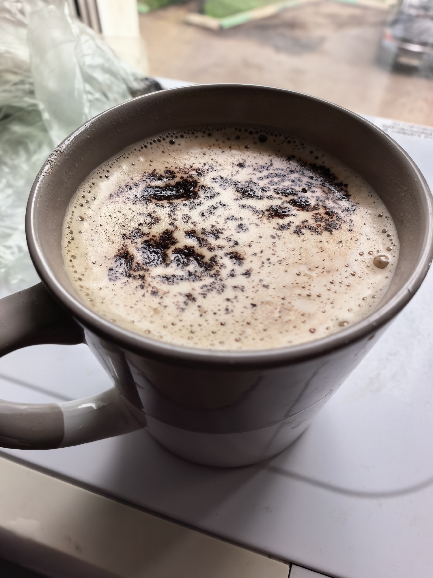 The image shows a close-up of a mug filled with coffee, topped with foam and sprinkled with what appears to be cocoa powder or cinnamon. The mug has a beige exterior with a lighter colored handle. In the background, out of focus, is a window showing an outdoor scene with a blurry view of a street, trees, and a parked car. To the left of the mug, a crumpled plastic bag is visible. The scene is set on a white surface, possibly a table or countertop, with a hint of another surface below. There is no indication of the location being in Sterlitamak, Russia.