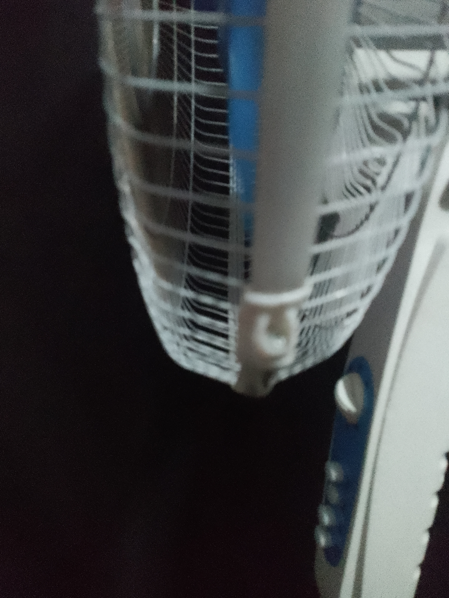 A close-up, low-angle shot shows the lower part of a white and blue oscillating fan. The fan cage, made of white plastic grid, is prominent in the foreground, with a blurred dark background. A small, white plastic piece is visible at the bottom of the fan cage, appearing to be a securing clip or support. To the right of the fan cage, the white plastic body of the fan is visible, with a blue accent strip and several blurry buttons or indicators. The image is out of focus, particularly the background. The overall impression is of a household appliance in an indoor setting.