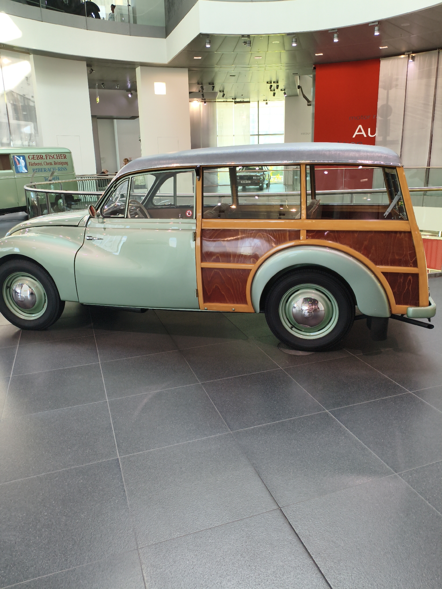 The image shows a vintage light green station wagon with wood paneling, likely an Audi 500, on display in a modern museum. The car is parked on a dark grey tiled floor. In the background, there is a red banner with white lettering that reads "motor r Au", suggesting this is the Audi Museum in Ingolstadt, Germany. The museum features modern architecture with high ceilings, glass railings, and spotlights. Other vehicles and displays are visible in the background, including a green van with text on its side. The lighting in the museum is bright and even, typical of an exhibition space.