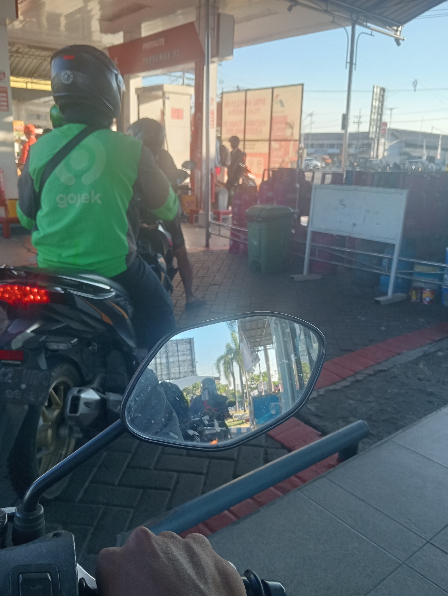 A first-person perspective shot captures a motorcycle rider at a gas station, likely in Sawotratap, Indonesia, based on the context provided by the clothing worn by one of the individuals. The rider, wearing a green Gojek jacket and a black helmet, is seated on a motorcycle. The motorcycle's rear is visible, including its red taillights. A side mirror reflects another motorcycle with a rider wearing a black helmet. In the background, there are gas pumps, a trash bin, and a white sign on a stand. The overall environment suggests a typical urban gas station setting.