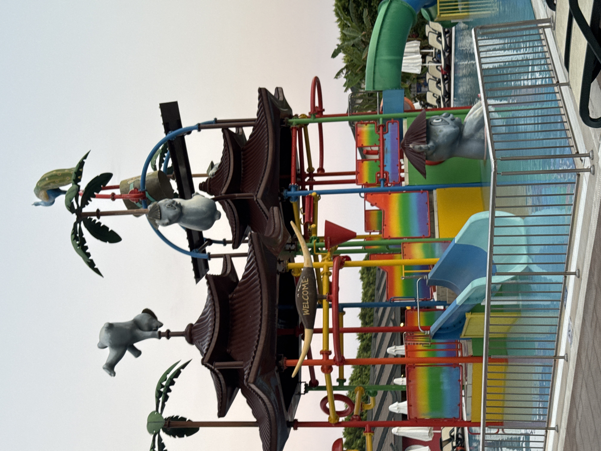 The image shows a water park playground with a central structure designed to resemble an Asian pagoda. The structure is adorned with colorful slides and animal figurines. In the foreground, a blue and teal slide enters a swimming pool. Several lounge chairs and umbrellas are visible in the background, as are lush green trees. The sky is overcast, suggesting it may be early morning or late afternoon. The scene is likely in Aksu, Turkey, a region known for its numerous resort hotels with water parks.