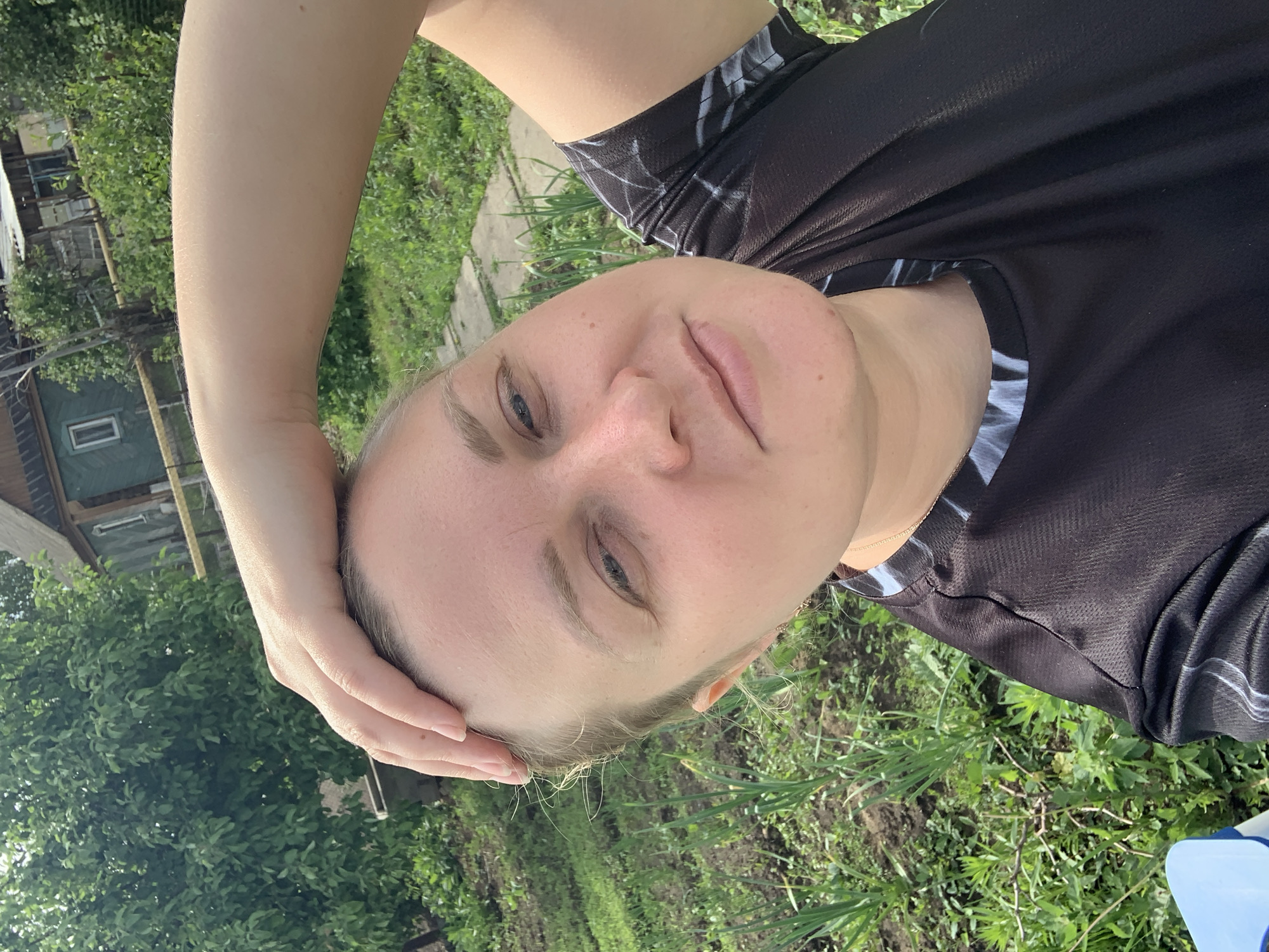 A woman with fair skin, light blonde hair, and blue eyes is positioned in the foreground of the image. She is wearing a black athletic shirt. Her left arm is raised and her hand is touching her forehead. The background includes a green lawn, trees, and a house with a visible window. The location is the city of Chaikovsky, Russia.
