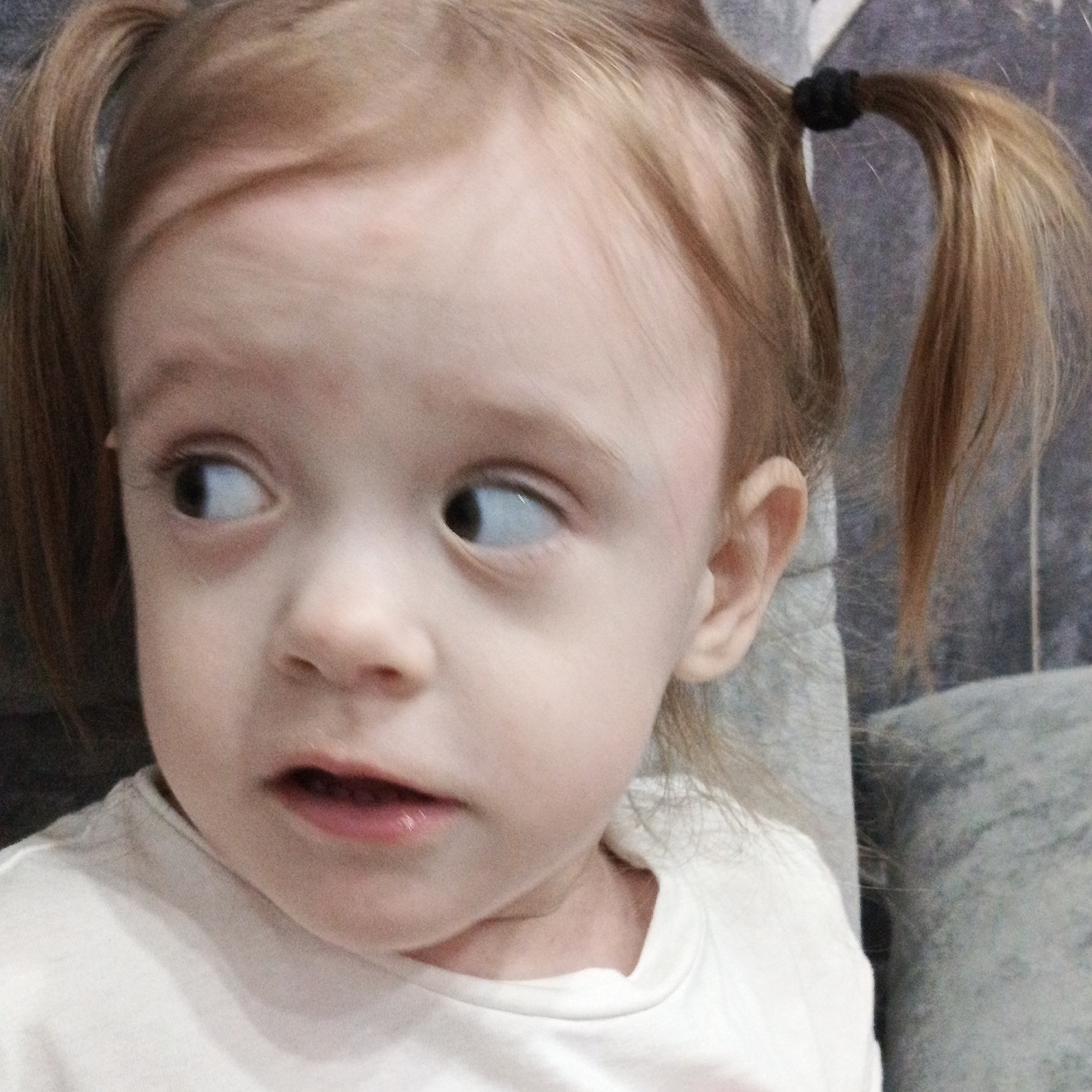 A young child with fair skin and reddish-blonde hair styled in pigtails is the subject of the image. The pigtails are secured with black hair ties. The child is wearing a light-colored t-shirt. The child is looking towards their left with a slightly open mouth, possibly in surprise or contemplation. The background appears to be a gray textured surface, possibly a wall or furniture. The location is Марковский, Russia.