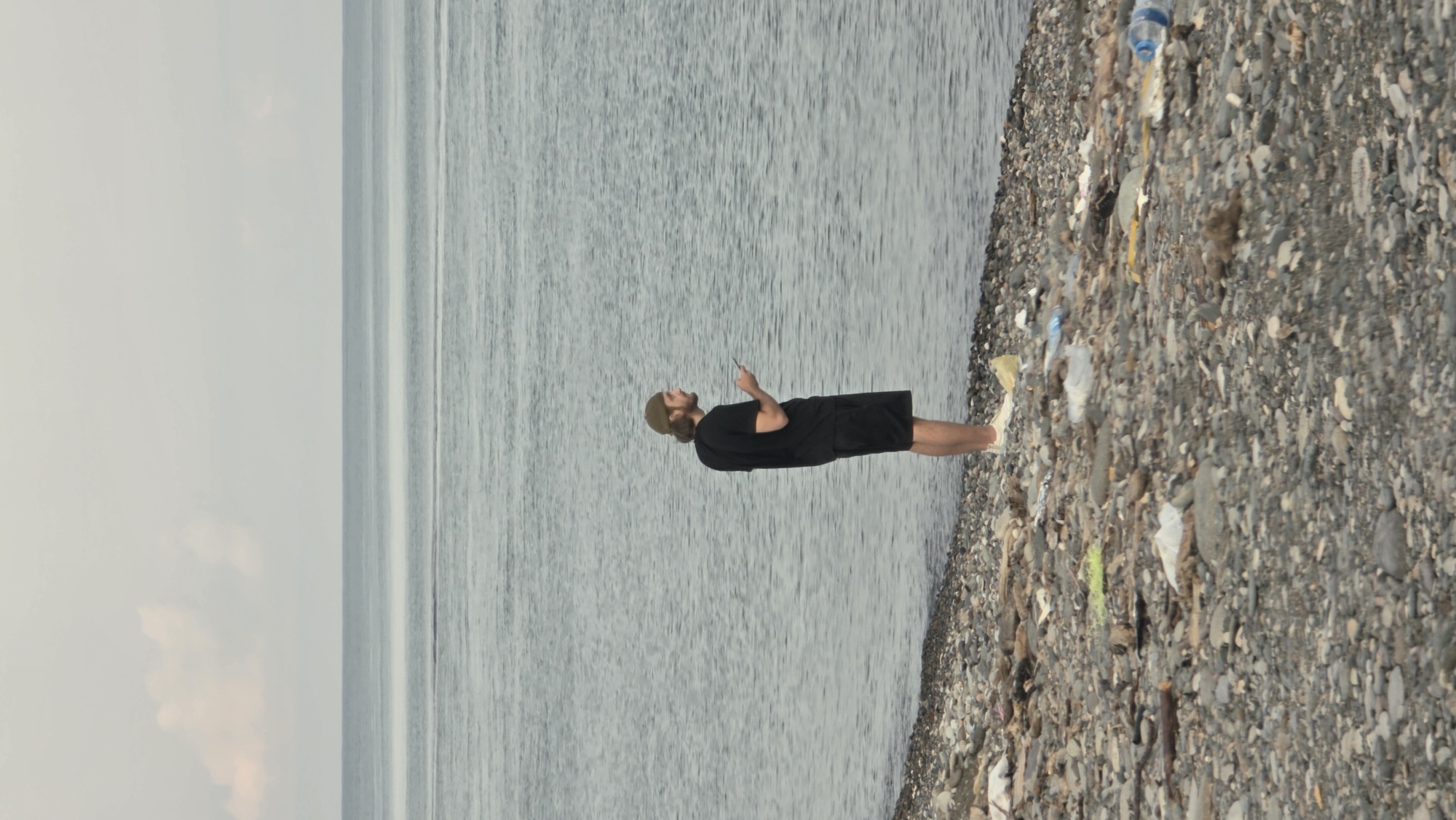 A man stands on a pebble beach strewn with litter, looking at his phone. He is wearing a black t-shirt, black shorts, a beige baseball cap, and white sneakers. The beach is next to a body of calm water, with a hazy horizon under a pale sky. The scene appears to be an outdoor environment with significant plastic and organic debris scattered across the shore.