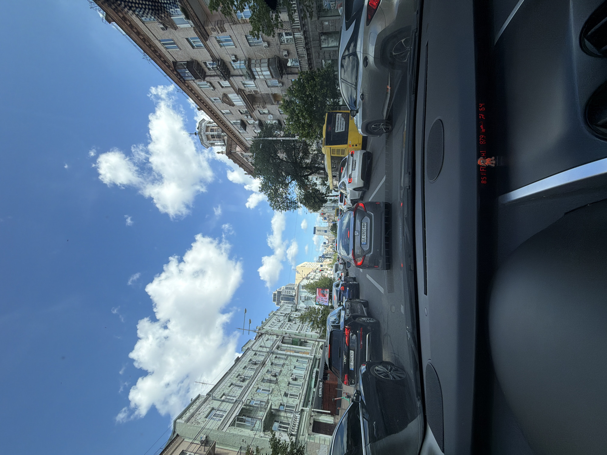 The image shows a view from inside a car driving in Kyiv, Ukraine, on a sunny day with scattered clouds. The car is in traffic on a multi-lane street lined with buildings. The architecture appears to be a mix of older, ornate buildings and more modern structures.
On the left, there is a light green building with decorative elements and balconies. Further down the street, more buildings with balconies are visible. On the right, there is a multi-story brick building with many windows and balconies. A yellow bus is stopped in traffic.
Several cars are visible in front of the vehicle from which the photo is taken, including a black Audi Q5, a dark grey Honda Civic, and a white Tesla. The dashboard of the car shows a digital display with what appears to be temperature readings and other vehicle information. A small figurine of a cartoon character is placed on the dashboard.
The street is paved and appears to be in good condition. Trees are planted along the sides of the street. The overall impression is that of a busy urban environment during daylight hours.