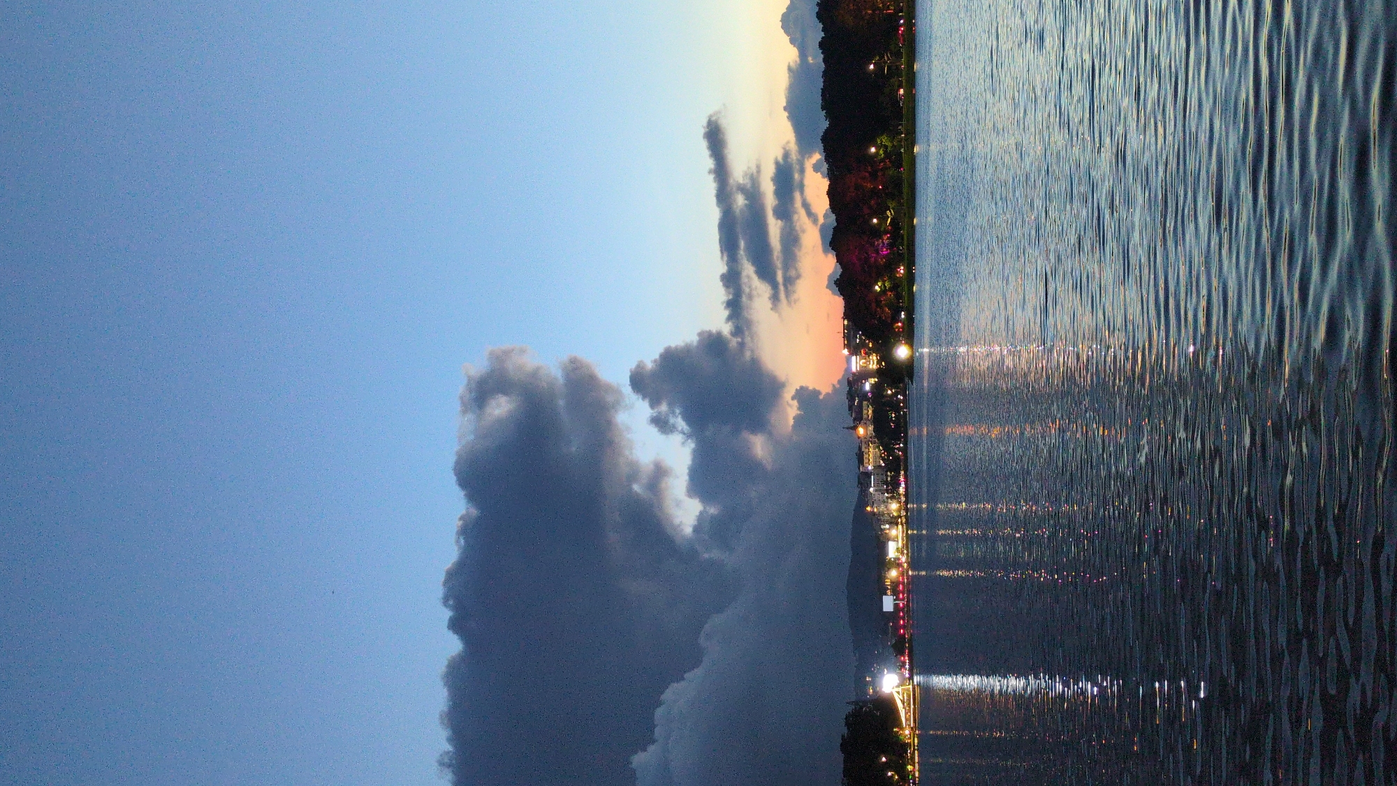 A twilight scene unfolds over Xuan Huong Lake in Da Lat, Vietnam. The sky, a gradient of deep blue to soft orange near the horizon, is dominated by a large, dark cloud formation on the left, contrasting with lighter clouds on the right. A distant cityscape with illuminated buildings and streetlights lines the far shore, their lights reflecting shimmering on the lake's surface. The water, rippling gently, captures the reflections of the lights, creating streaks of white and color. Trees and hills form a dark silhouette against the illuminated city. The overall impression is a serene and atmospheric urban landscape at dusk.