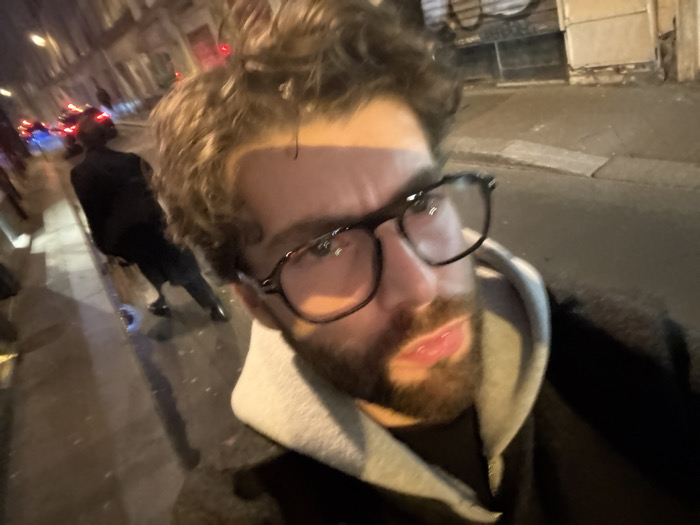 The image presents a nighttime urban street scene, likely in Paris, France, captured from a low-angle selfie perspective. In the foreground, a male individual with curly brown hair, a beard, and dark-framed glasses looks directly at the camera with a neutral expression, his mouth slightly open. He is dressed in a dark jacket over a grey hooded sweatshirt. His face is illuminated from the right, casting shadows on the left.
Behind the primary subject, to the left, another individual in dark attire is walking away from the camera on a wet, reflective sidewalk. This person appears to be handling a small, light-colored object near their right leg, possibly a kick scooter or a package. Further down the street, multiple vehicles are visible, displaying red taillights and some blue/white headlights, indicating ongoing traffic. The street surface itself appears dark and wet, reflecting ambient light.
The background consists of multi-story buildings consistent with Parisian architecture, featuring stone facades and numerous windows, lining both sides of the thoroughfare. Some ground-level establishments appear to have closed shutters or boarded entrances. The scene is illuminated by artificial streetlights and vehicle lights, creating reflections on the damp ground. The overall architectural style, street layout, and urban density are consistent with a street within Paris.