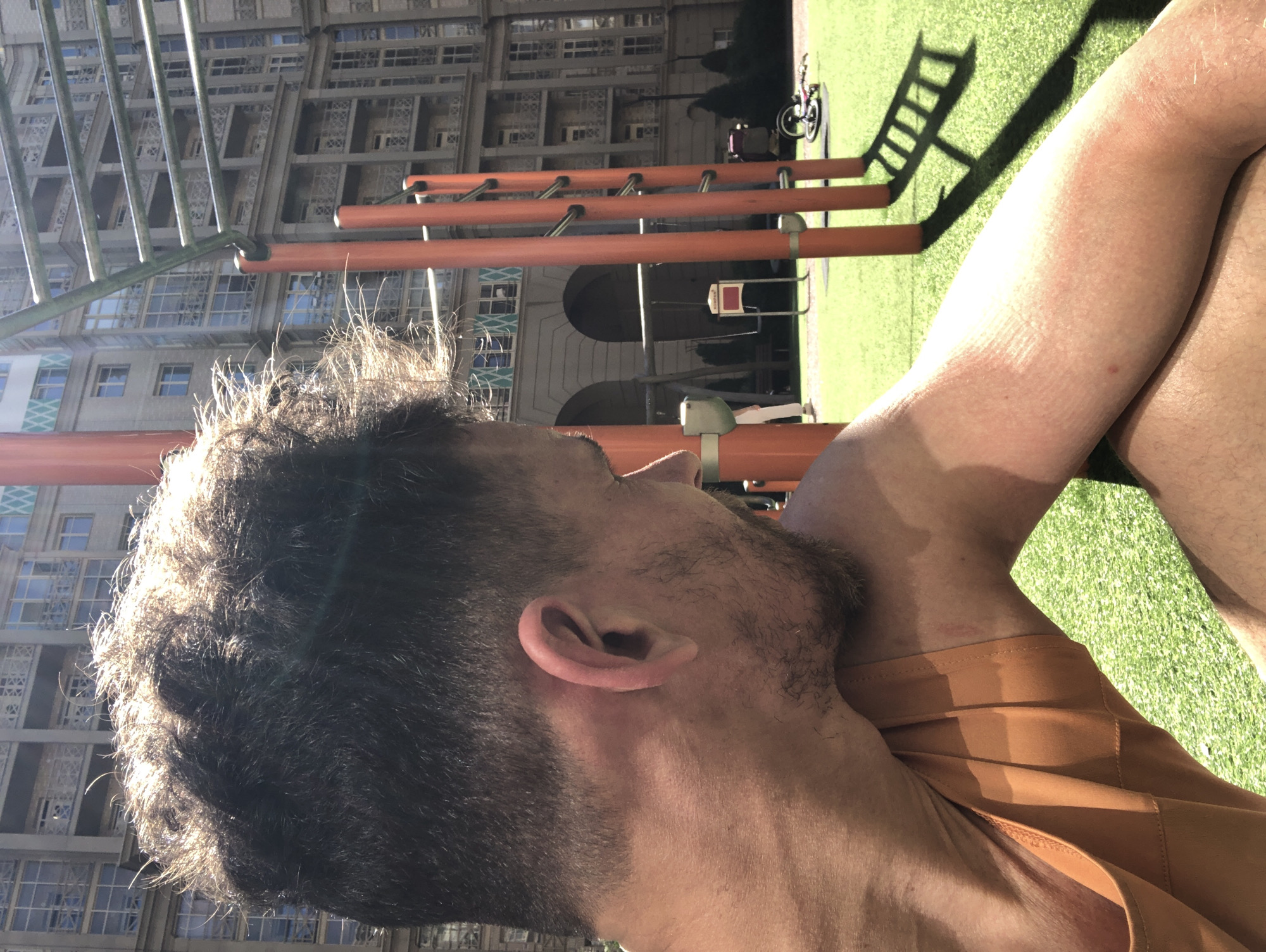 An outdoor scene in Saint Petersburg, Russia, features a male individual in the foreground, viewed from the mid-back and right profile, seated or crouched on artificial green turf under direct sunlight. He possesses dark, disheveled hair strongly backlit, creating a halo effect, and light stubble on his jawline. He wears an orange tank top, and his exposed skin exhibits some redness. A faint red mark is visible on his upper left arm/shoulder area. His gaze is directed downwards towards his right arm. In the midground, prominent orange and silver metal outdoor exercise equipment, resembling monkey bars, stands on the turf, casting long, sharp shadows. To the left of the exercise equipment, a swing set and a partially visible white sign with red text ("KOMPAKT") are present. To the far right, a child's bicycle lies on its side on the green surface. The background is dominated by large, multi-story residential buildings with numerous windows, balconies, and classical architectural details, including arched ground-level entrances on one structure and light green diamond patterns near the roofline of another. The sky is clear, indicating bright, sunny weather.