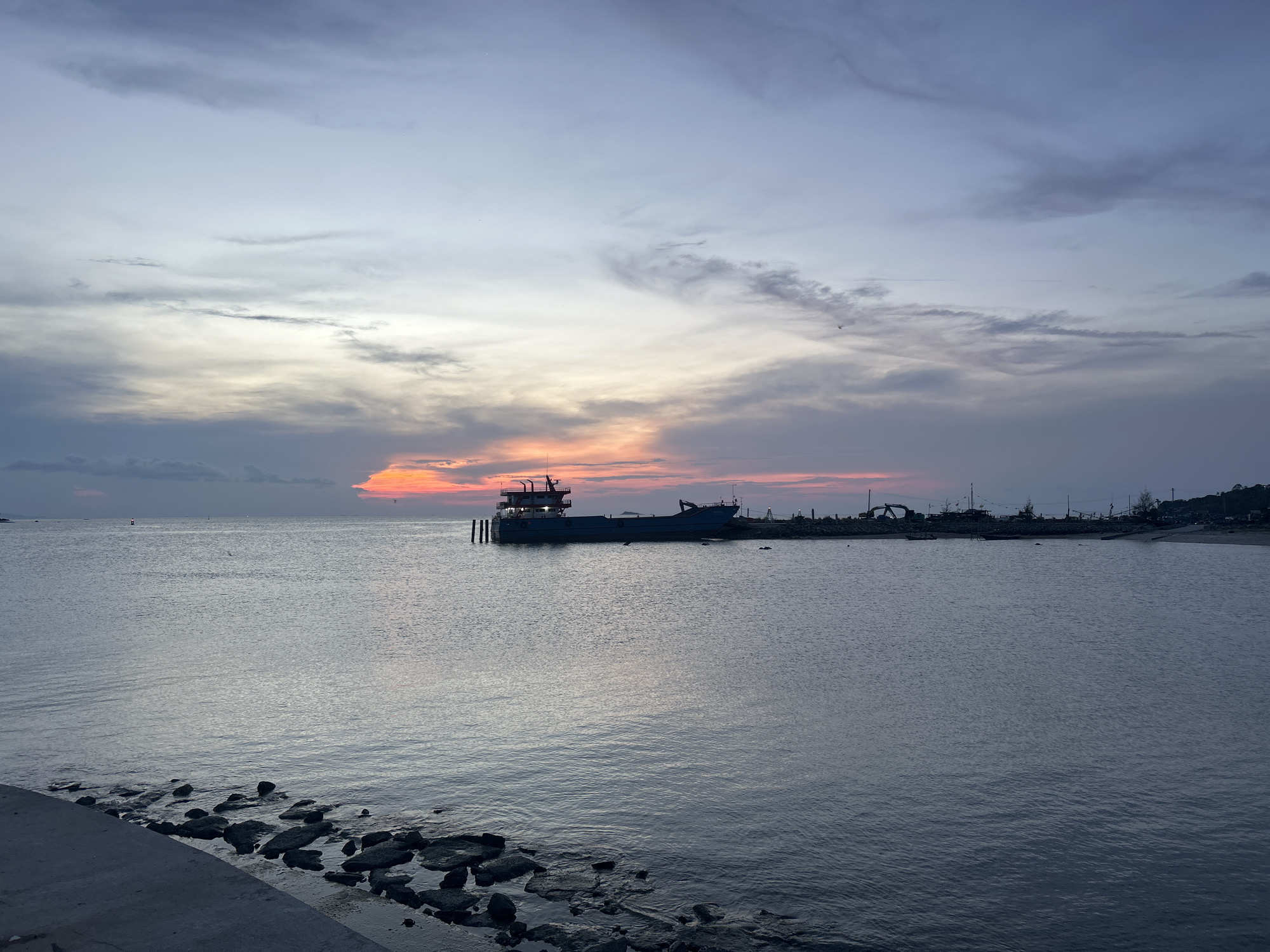 The image depicts a dusk scene in Thong Sala, Thailand. A large barge is docked offshore, with its lights illuminated against the fading orange glow of the setting sun. The sea is calm, with subtle ripples reflecting the sky's colors. The foreground shows a rocky shoreline transitioning into a concrete embankment. In the distance, the coastline of Thong Sala is visible with sparse vegetation and structures. The sky is filled with a mix of grey and orange clouds.