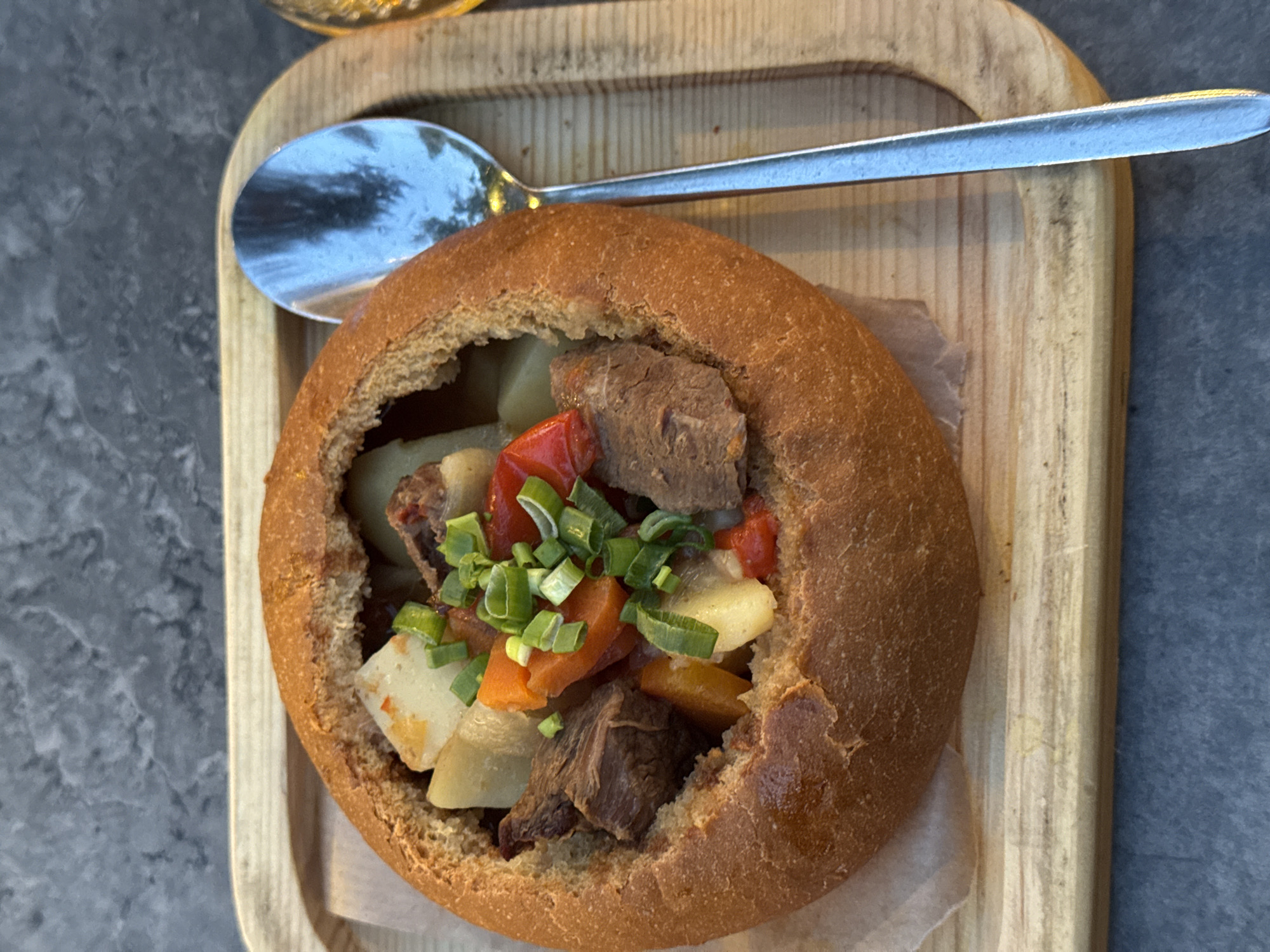The image captures a close-up, overhead view of a hearty stew served in a rustic bread bowl. The main subject is a round, golden-brown bread loaf, hollowed out to create a bowl. Its crust is visibly textured and robust.
Inside the bread bowl, the stew is generously filled with substantial chunks of dark brown meat, likely beef, alongside diced white potatoes, bright orange carrots, and vibrant red bell peppers. The dish is garnished with a generous scattering of freshly chopped green onions, adding a fresh, green accent.
The bread bowl sits on a piece of light brown parchment paper, which in turn rests on a light-colored wooden serving tray. The tray has a noticeable wood grain and a slightly raised, rounded rim on its edges. A silver spoon with a reflective surface is positioned on the wooden tray, to the left of the bread bowl. The spoon reflects blurry images, possibly of the ceiling or immediate surroundings.
The wooden tray is placed on a dark gray, subtly textured surface, resembling stone or marble. In the upper left corner of the frame, a blurred, yellowish-amber object, potentially a glass of drink, is partially visible. The lighting suggests an indoor setting, likely for a meal. No specific activity beyond the presentation of food is depicted, and no visible text is present. The dish appears warm and comforting, characteristic of meals found in regions like Izhevsk, Russia.