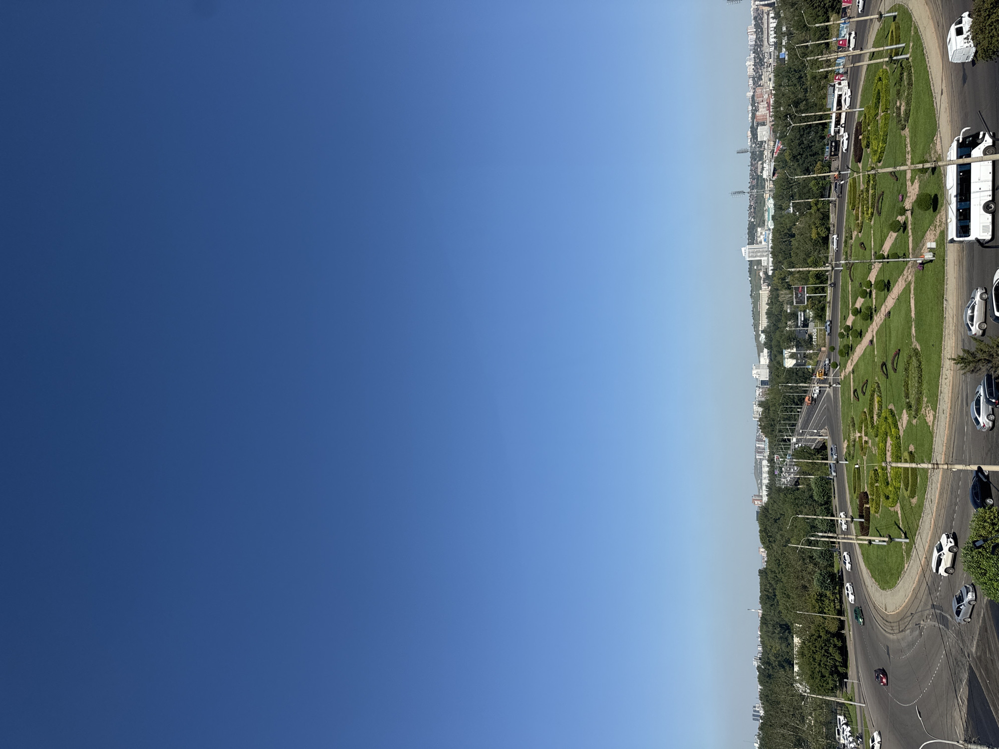 This is a high-angle, wide shot of a busy city intersection in Krasnoyarsk, Russia, on a clear, sunny day. The sky is a vibrant, cloudless blue, dominating the upper portion of the frame. Below, a complex road network with multiple lanes curves and converges. A large, circular green space with manicured hedges and trees sits at the heart of the intersection, acting as a traffic island. Several vehicles, including cars and buses, are captured in motion on the roads. In the background, a dense line of trees forms a natural barrier, beyond which a cityscape unfolds with various buildings and structures, suggesting a developed urban area. The overall scene depicts a typical urban environment with traffic and greenery.
