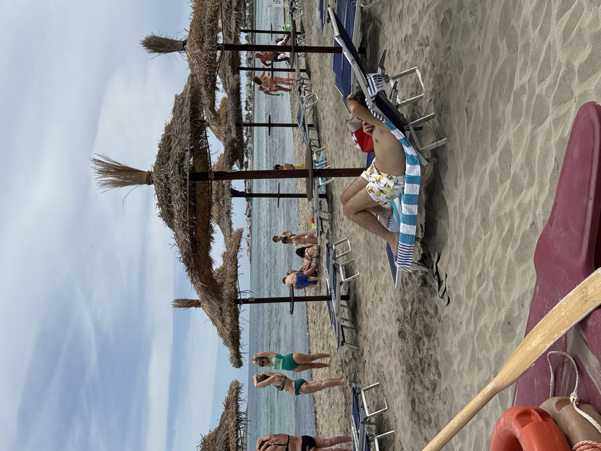 The image depicts a crowded beach in Isola delle Femmine, Italy, under a cloudy sky. In the foreground, a wooden oar rests on a maroon floatation device, suggesting some water activity is present or imminent. The beach is covered in light-colored sand and lined with numerous straw-thatched umbrellas and lounge chairs.
Several people are visible on the beach. In the mid-ground, a man in patterned swim trunks is lying on a lounge chair, his eyes closed and hands behind his head, appearing to be resting or napping. Further back, a group of people are standing near the water's edge. Two women in swimsuits are facing the water, one in a green one-piece and the other in a green and black bikini. Behind them, a man in blue shorts is sitting on the sand with a child. To the right, a group of children are playing near the water. Further in the background, more beachgoers are visible, some walking along the shore and others under umbrellas. The sea is a calm, blue-green color, with a distant coastline visible. The overall scene conveys a typical summer day at a popular beach resort.