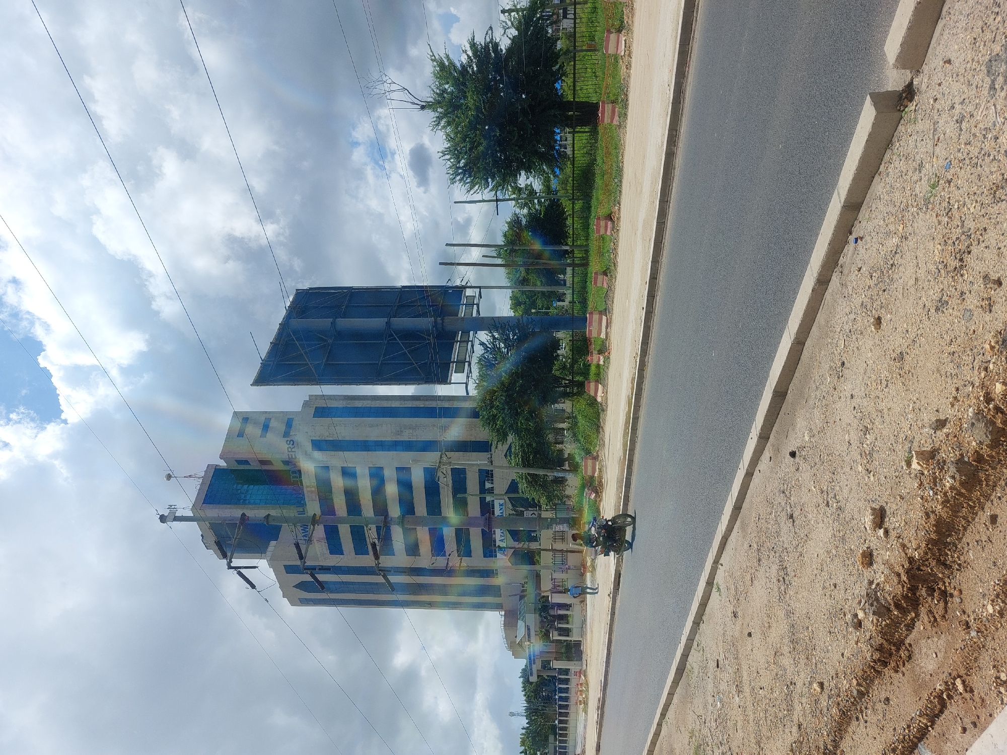 The image shows a view of the Azura Towers building in Dar es-Salaam, Tanzania, alongside a road and sidewalk. The towers are a modern, high-rise structure with blue glass windows. An empty billboard is positioned next to the building. A motorcycle is driving on the road. Vegetation, including trees and grass, lines the sidewalk. Overcast skies are visible in the background.