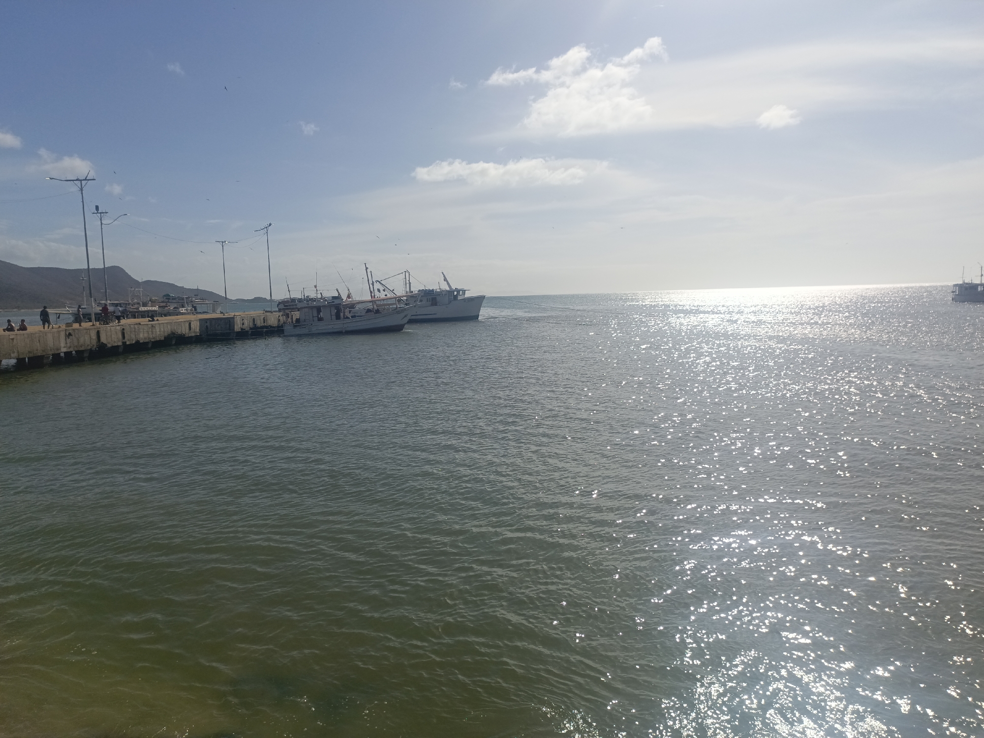 The image depicts a coastal scene in Juan Griego, Venezuela. A concrete pier extends into the water from the left. People are present on the pier. Two boats are docked alongside it. A body of water reflects sunlight. In the background, land and mountains are visible. The sky is blue with some white clouds. Utility poles line the pier.