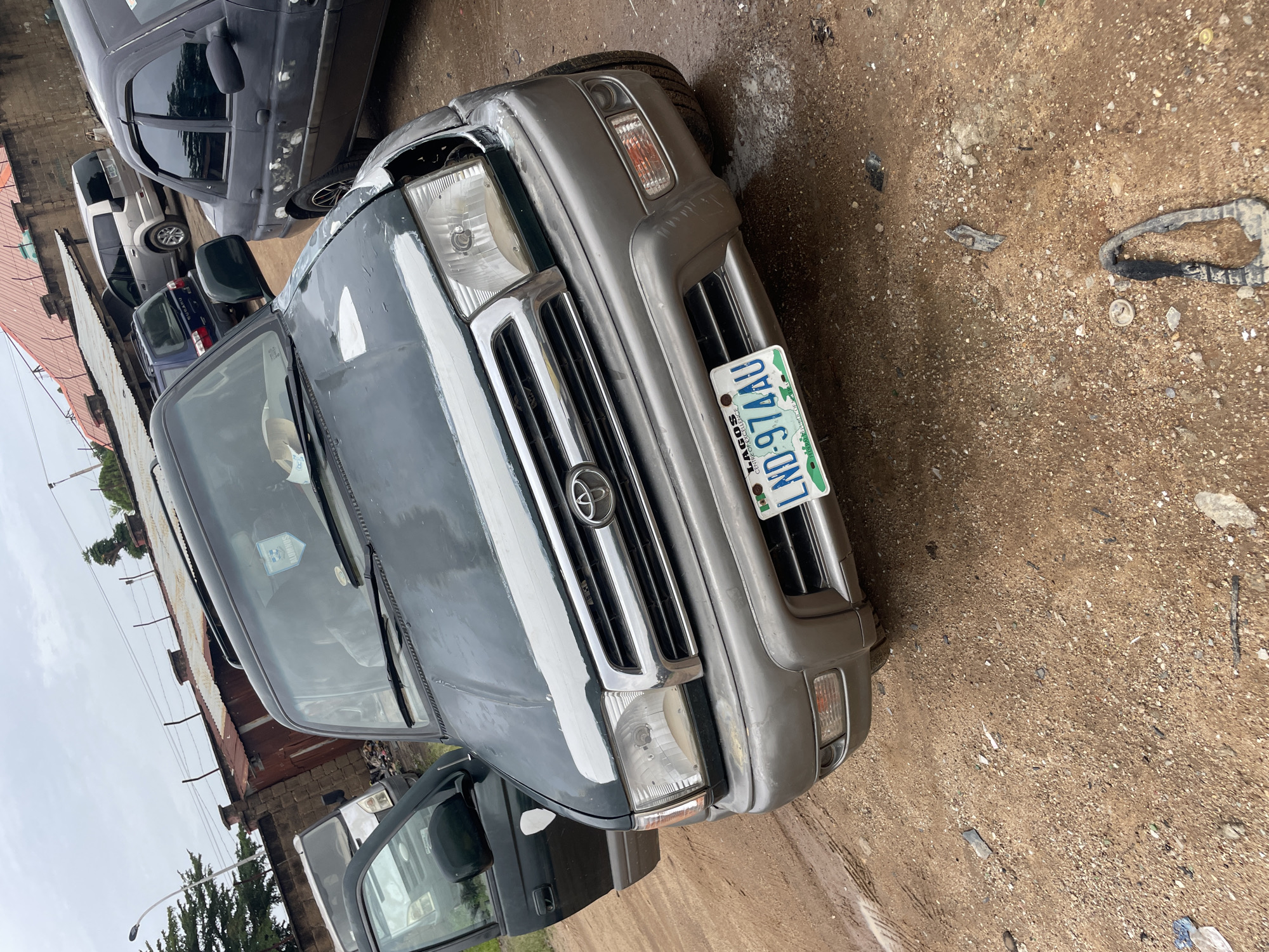 The image shows a dark green Toyota 4Runner parked on a dirt lot in an outdoor setting. The vehicle is facing forward, with its driver's side door open. The front of the car shows some damage, including a broken headlight and chipped paint on the bumper and hood. A license plate with the text "LAGOS LND-974AU" is visible on the front bumper. In the background, other vehicles and a corrugated metal structure are present. The ground is covered in dirt and gravel, with some debris scattered around. The sky is overcast.