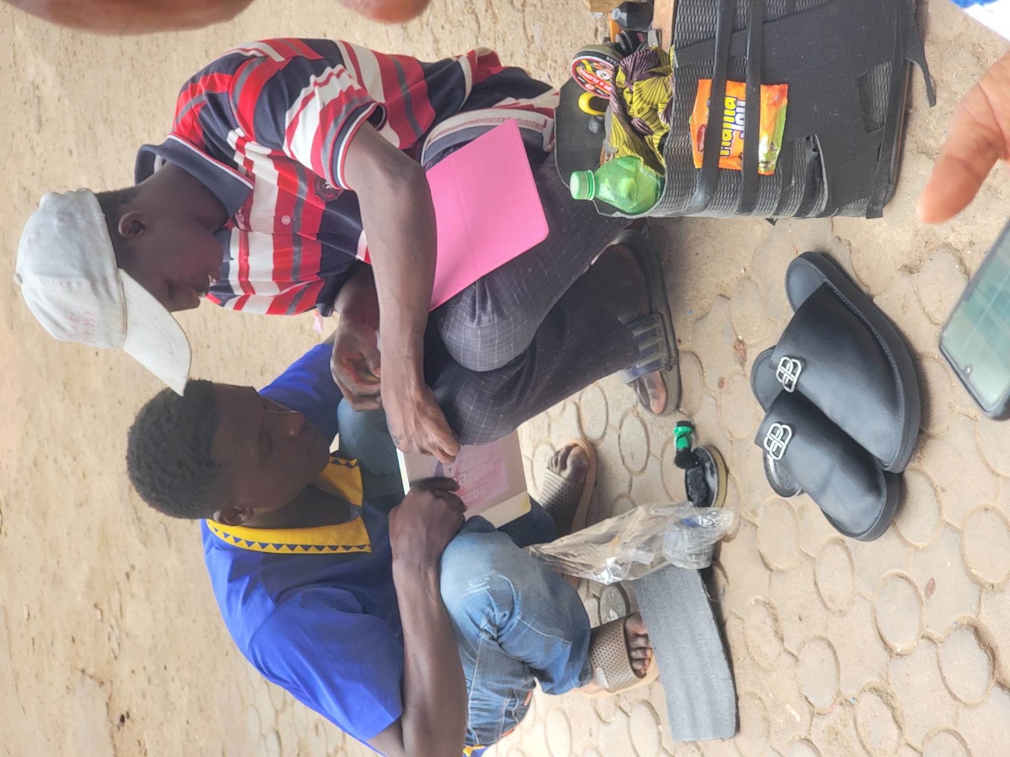 Two men are crouched close together on a paved surface. One man wears a white baseball cap, a red, white, and blue striped shirt, and patterned pants. The other man wears a blue shirt with a yellow triangular pattern around the collar and blue jeans. They both are looking at a pink folder/book. To their right is a black suitcase that has some snacks on top, a green water bottle, a pair of black slippers with a silver logo on them, and a mobile phone. The pavement beneath them appears to be patterned with interconnected circular shapes. The scene appears to be taking place outdoors. Location: Jos, Nigeria.